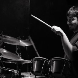 Vitalii Ishchenko - Czarno-białe zdjęcie perkusisty z uśmiechem, trzymającego pałeczkę nad zestawem perkusyjnym. Ujęcie z boku, ciemne tło, widoczne talerze i bębny.