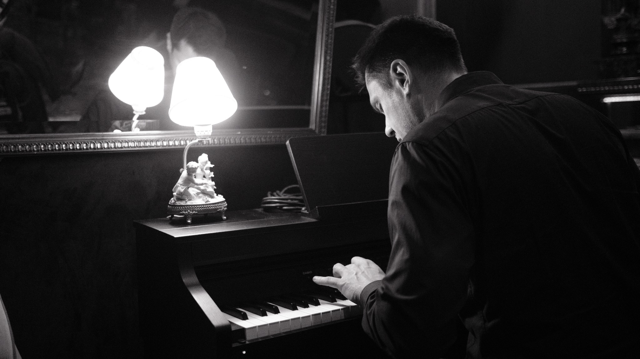 Czarno-białe zdjęcie mężczyzny grającego na pianinie w pokoju z lampką nocną i lustrem w tle. Ujęcie zza pleców, skupione na dłoniach na klawiszach.