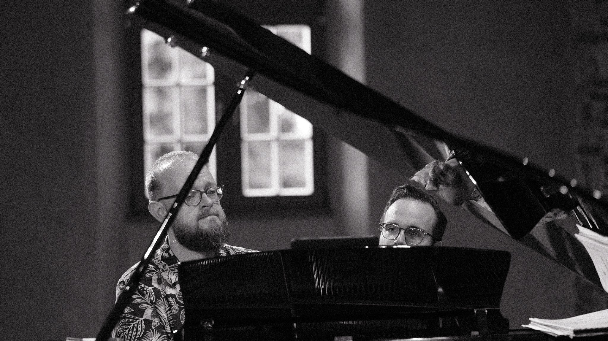 Czarno-białe zdjęcie dwóch mężczyzn w okularach grających na fortepianie. Jeden w kwiecistej koszuli, drugi zza instrumentu. Scena z koncertu lub sesji nagraniowej.