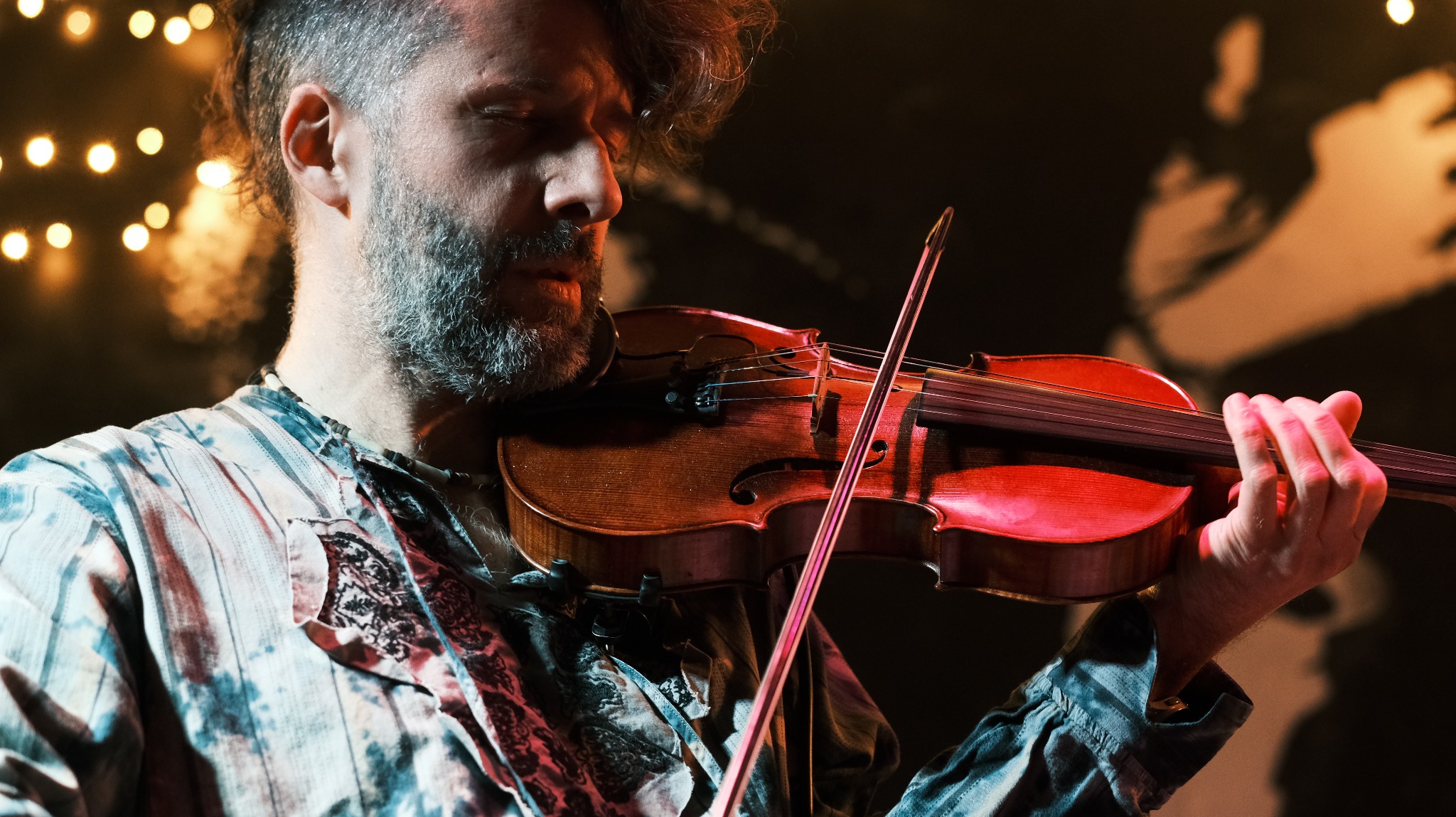 Mężczyzna z brodą gra na skrzypcach w skupieniu, oświetlony ciepłym światłem. W tle rozmyte światła i sylwetka postaci. Ujęcie z bliska, podkreślające emocje muzyka.