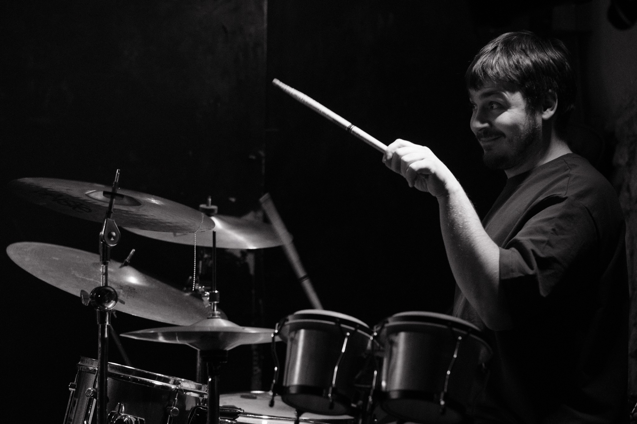 Czarno-białe zdjęcie perkusisty z uśmiechem, trzymającego pałeczkę nad zestawem perkusyjnym. Ujęcie z boku, ciemne tło, widoczne talerze i bębny.