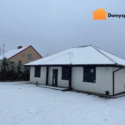 Domy z paczki. pl - Zimowy krajobraz z białym, parterowym domem z czarnymi akcentami i śnieżnym dachem. W tle inny dom i drzewa, wszystko pod delikatnym opadem śniegu.