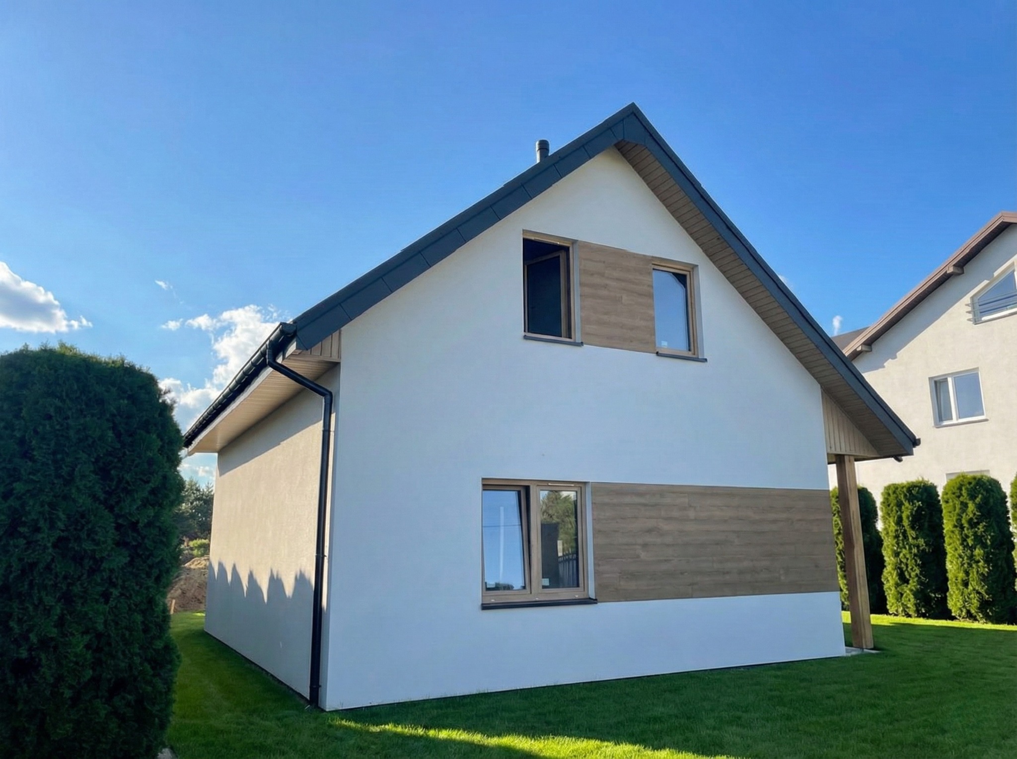 Gotowy dom z białymi ścianami i drewnianymi akcentami, widoczny z zewnątrz na tle zielonego trawnika i błękitnego nieba. Otwarta górna okiennica dodaje unikalności.