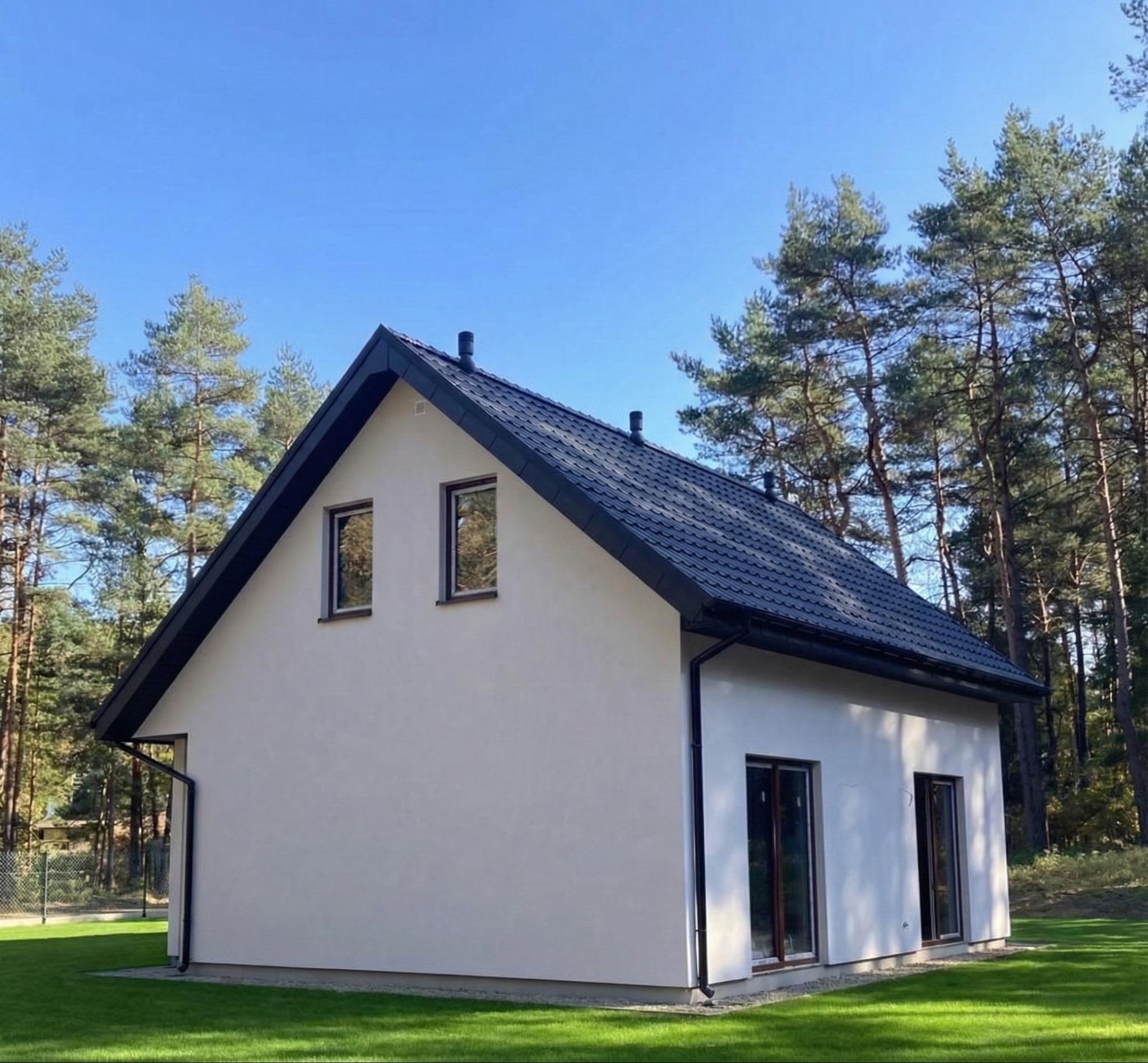Boczny widok na parterowy dom z ciemnym dachem na tle lasu i błękitnego nieba. Minimalistyczna bryła, jasna elewacja, ciemne okna i rynny. Zadbane otoczenie.