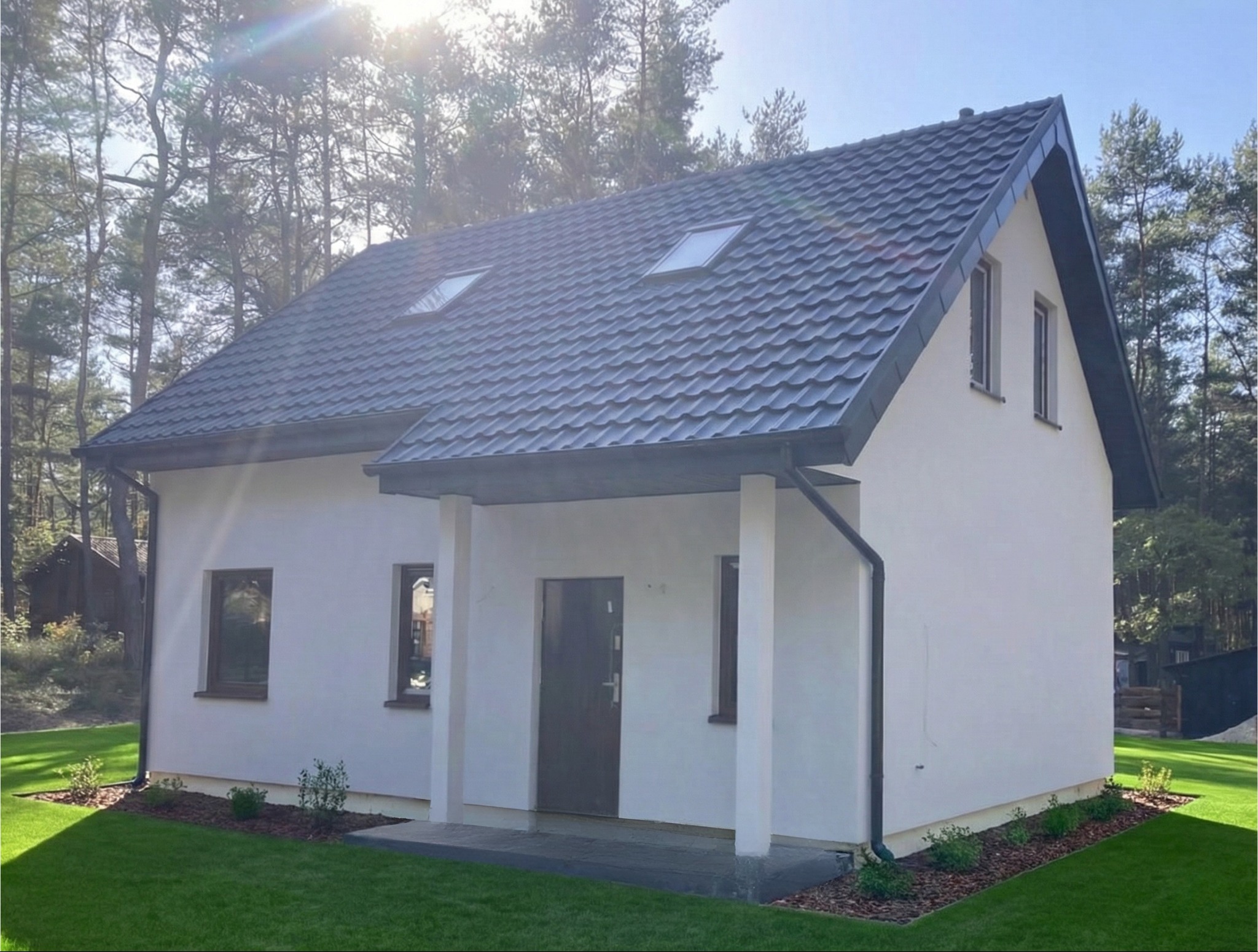 Gotowy, parterowy dom z ciemnoszarą dachówką, białymi ścianami i zadaszonym wejściem, otoczony zielonym trawnikiem i drzewami. Słoneczny dzień.