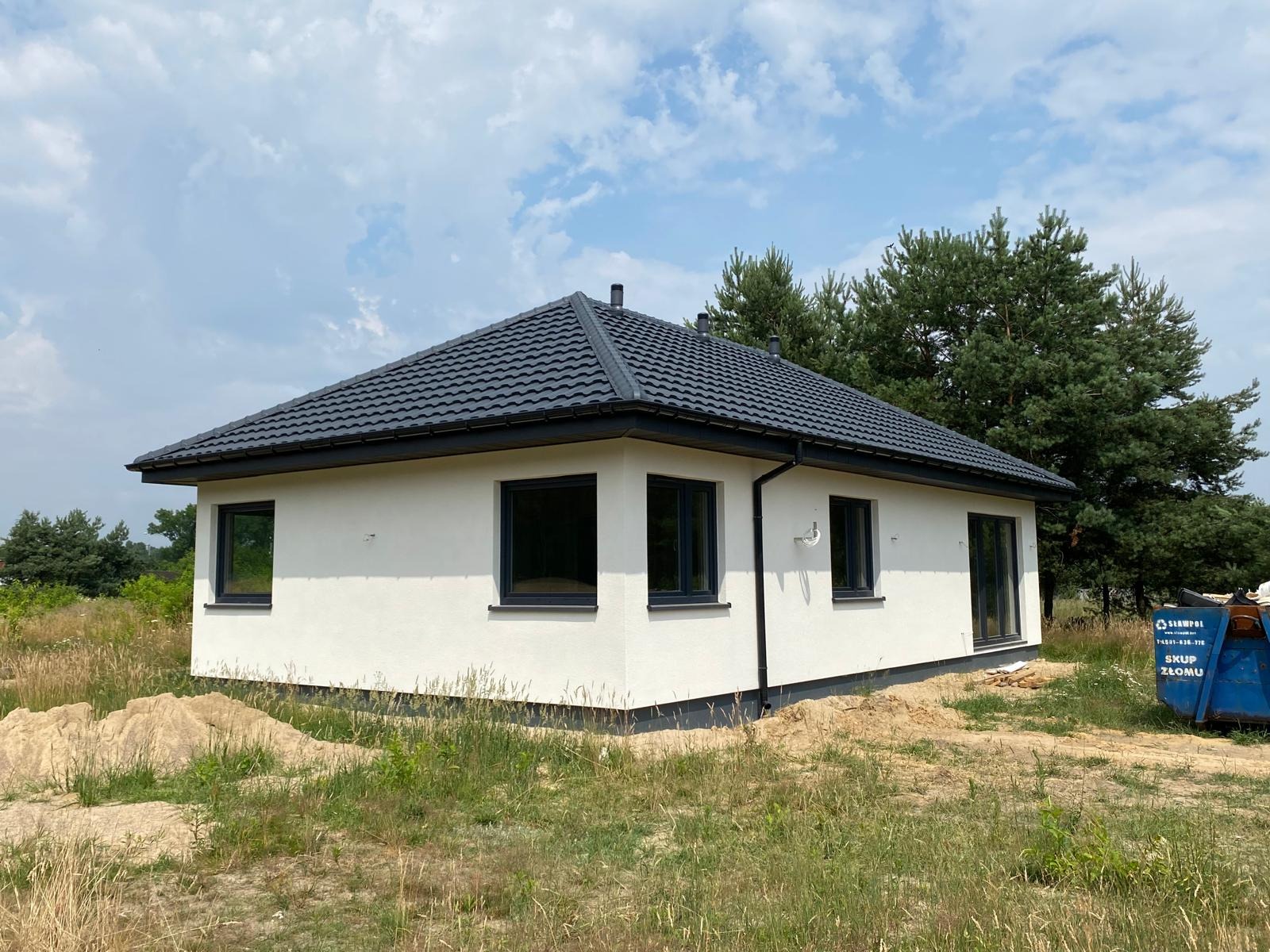 Gotowy, parterowy dom z ciemną dachówką i ramami okiennymi, otoczony zielenią i piaskiem. W tle widoczne drzewa i błękitne niebo z chmurami.
