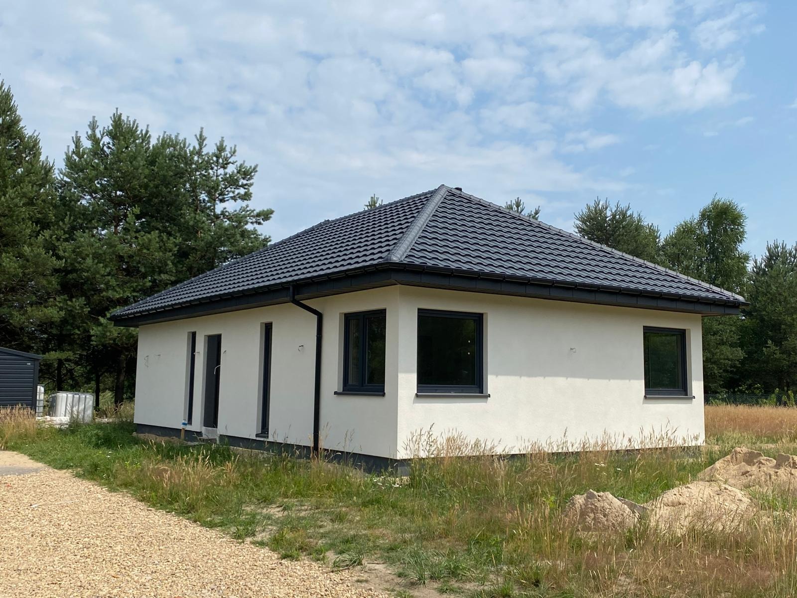 Gotowy, parterowy dom z ciemną dachówką i grafitową stolarką okienną, otoczony zielenią. Widok z zewnątrz, w tle drzewa i błękitne niebo.