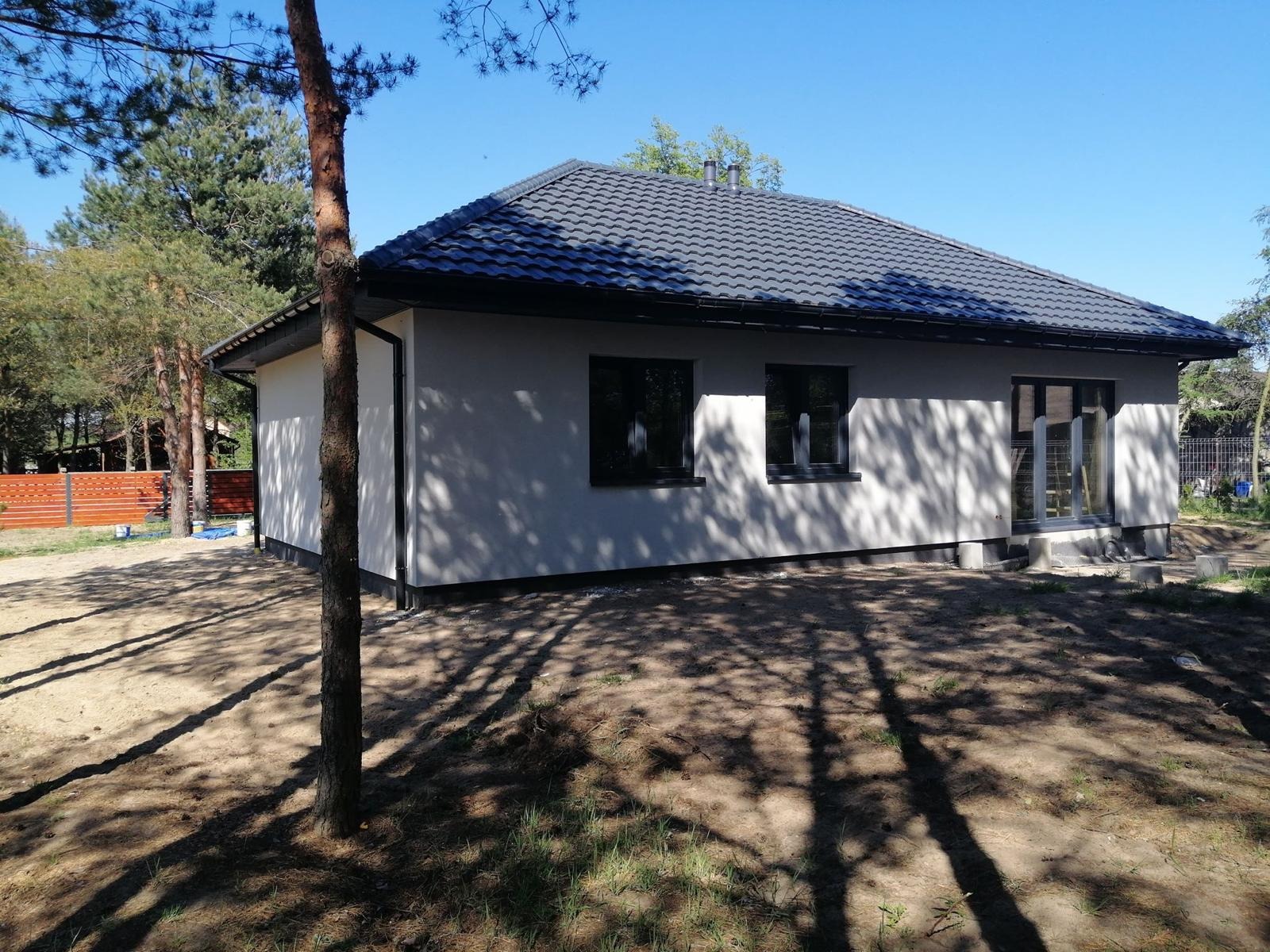 Parterowy dom z szarą dachówką i jasną elewacją, widziany z zewnątrz w słoneczny dzień. Cień drzewa na fasadzie. Widoczne okna i drzwi balkonowe.