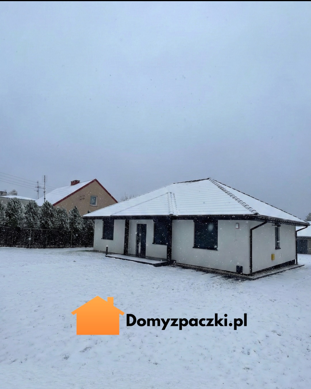 Zimowy krajobraz z białym, parterowym domem z ciemnymi obramowaniami okien i dachu, pokrytego śniegiem. W tle widoczny inny budynek i drzewa.