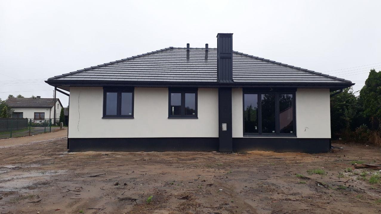 Parterowy dom z ciemną dachówką i stolarką okienną, elewacja w jasnym odcieniu. Widoczne elementy wykończeniowe i otoczenie w trakcie prac.