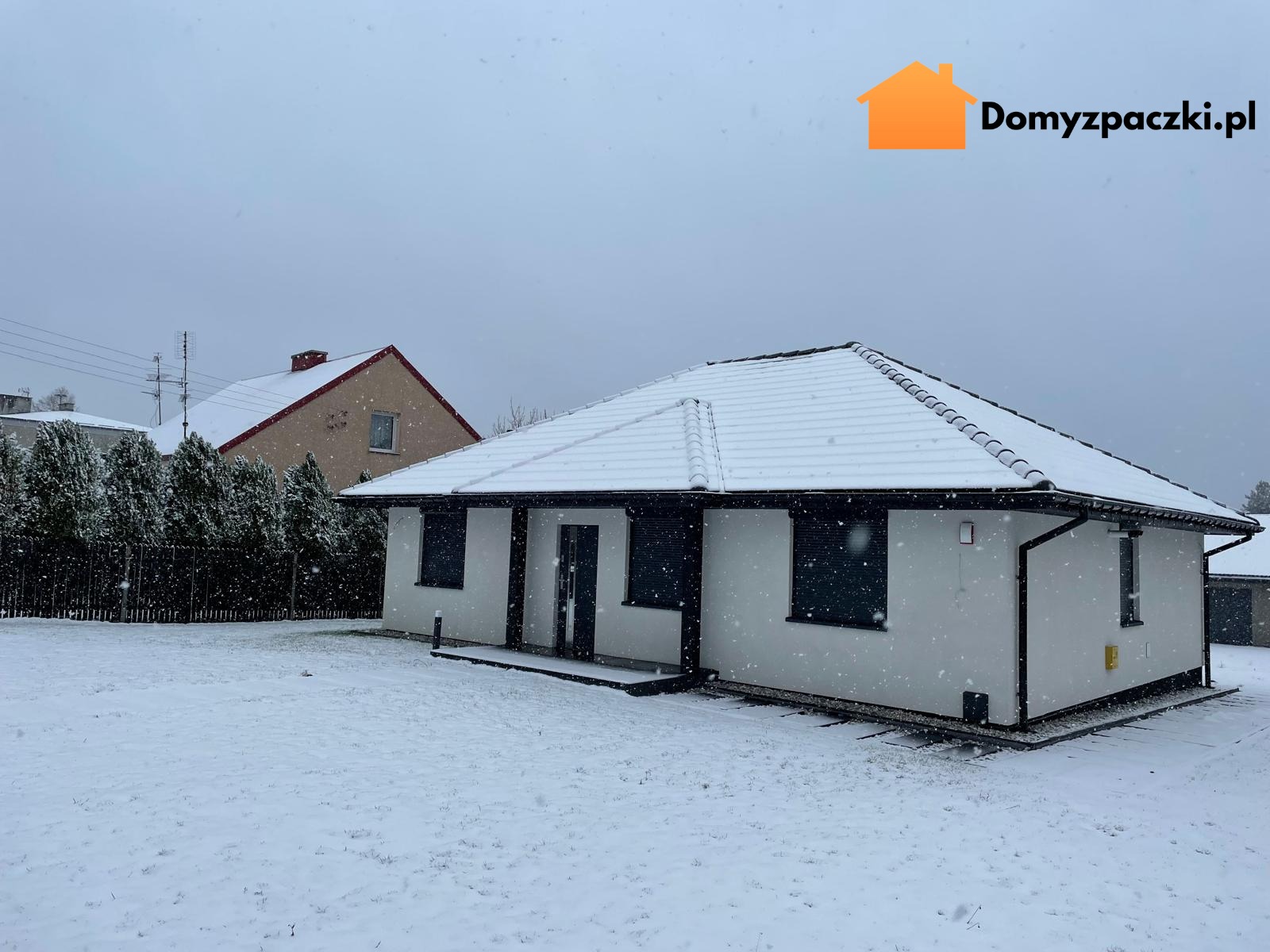 Zimowy krajobraz z białym, parterowym domem z czarnymi akcentami i śnieżnym dachem. W tle inny dom i drzewa, wszystko pod delikatnym opadem śniegu.