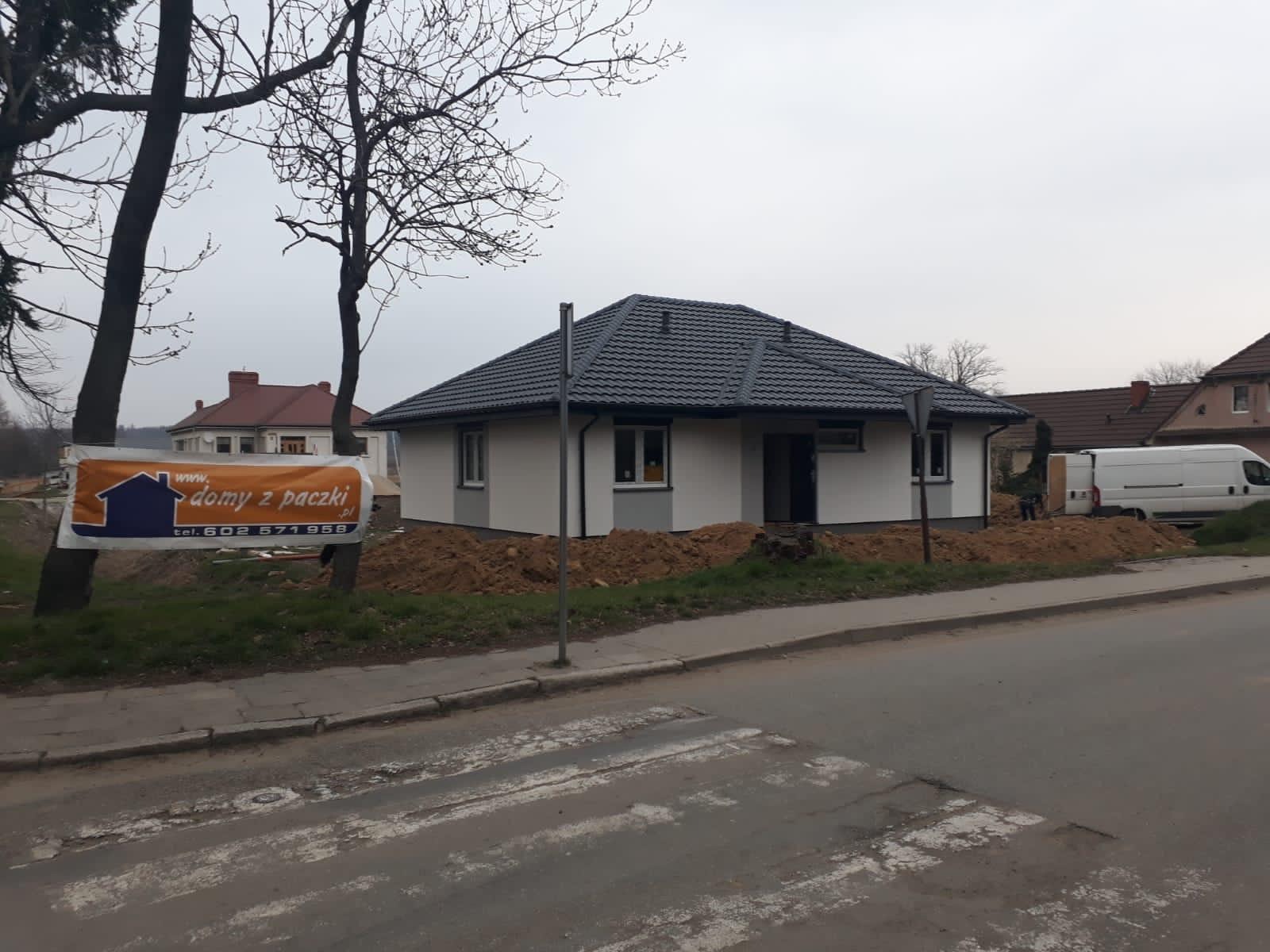 Niedokończony dom parterowy z ciemną dachówką, widoczny z ulicy z przejściem dla pieszych. Na pierwszym planie baner reklamowy firmy budowlanej.