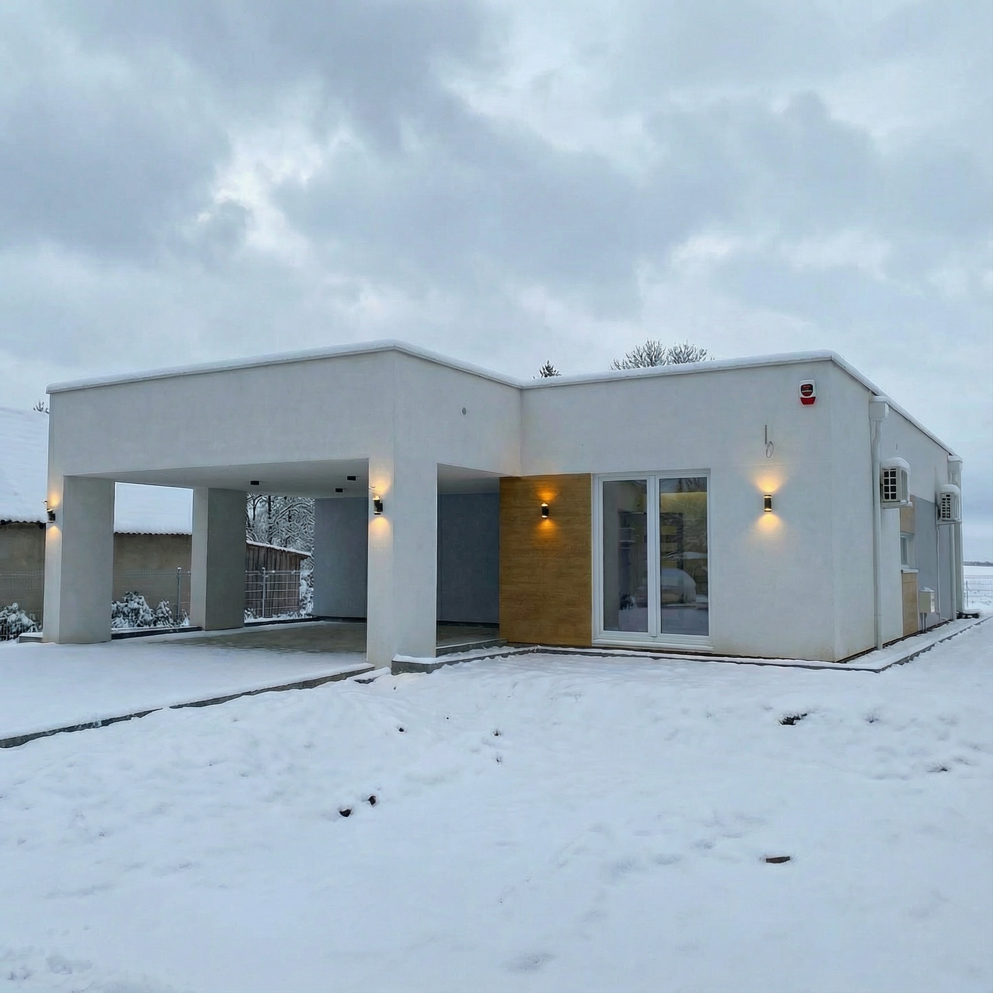 Nowoczesny, parterowy dom modułowy z płaskim dachem i wiatą garażową, oświetlony lampami elewacyjnymi, w zimowej scenerii. Widoczny system alarmowy i klimatyzacja.