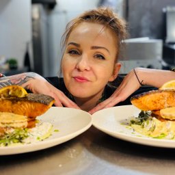 Restauracja Fantazja - Uśmiechnięta kobieta z tatuażami prezentuje dwa talerze z pieczonym łososiem i cytryną na tle profesjonalnej kuchni. Danie gotowe do serwowania.
