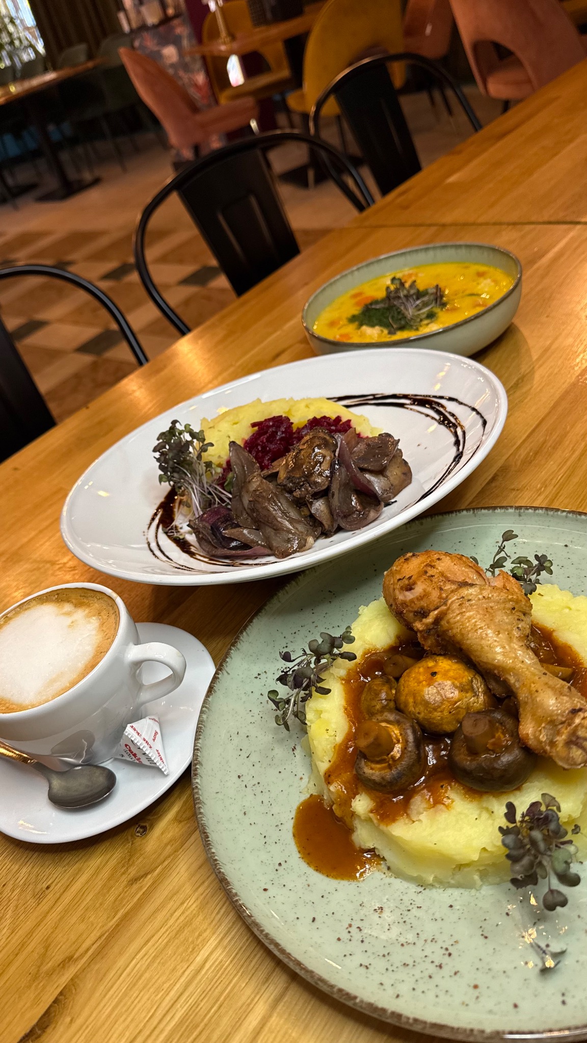 Zestaw obiadowy: noga kurczaka z puree i grzybami, wątróbka z puree i buraczkami, zupa krem oraz kawa cappuccino na drewnianym stole w restauracji.