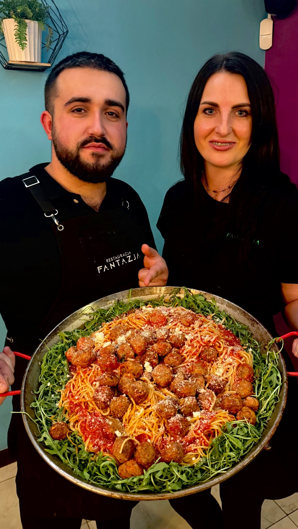 Dwoje pracowników restauracji 'Fantazja' prezentuje ogromną porcję spaghetti z klopsikami w metalowej patelni, otoczonej rukolą. Danie posypane parmezanem, na tle turkusowej ściany.