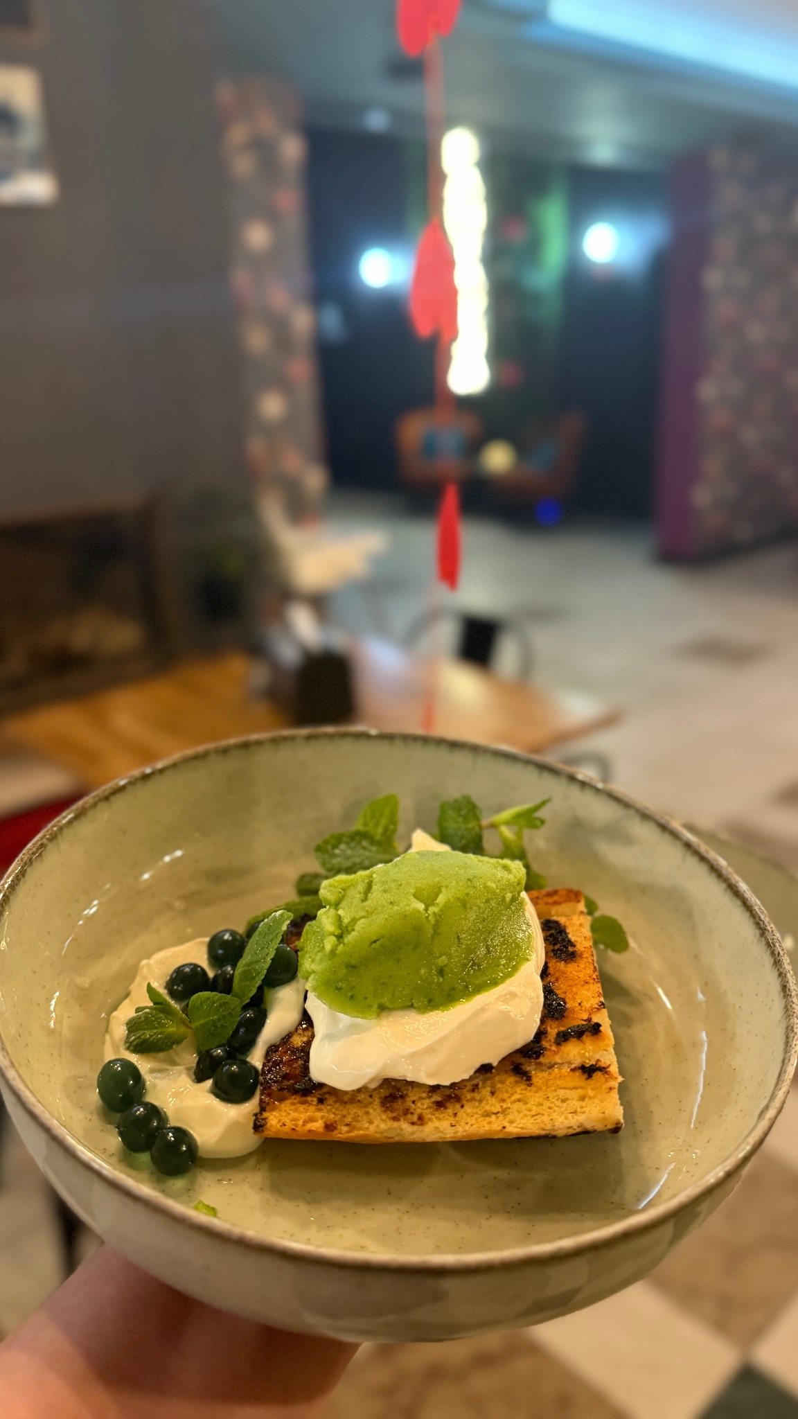 Apetyczna kompozycja: tost z kremem, zielonym musem i borówkami w ceramicznej miseczce, na rozmytym tle wnętrza restauracji z dekoracjami.