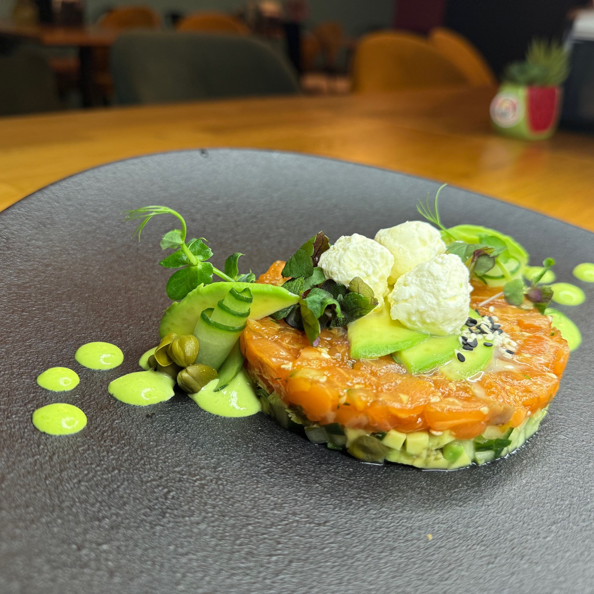 Tartar z łososia z awokado, kaparami i kremowym serem, udekorowany zieleniną i sosem, podany na czarnym talerzu. Elegancka kompozycja kulinarna.