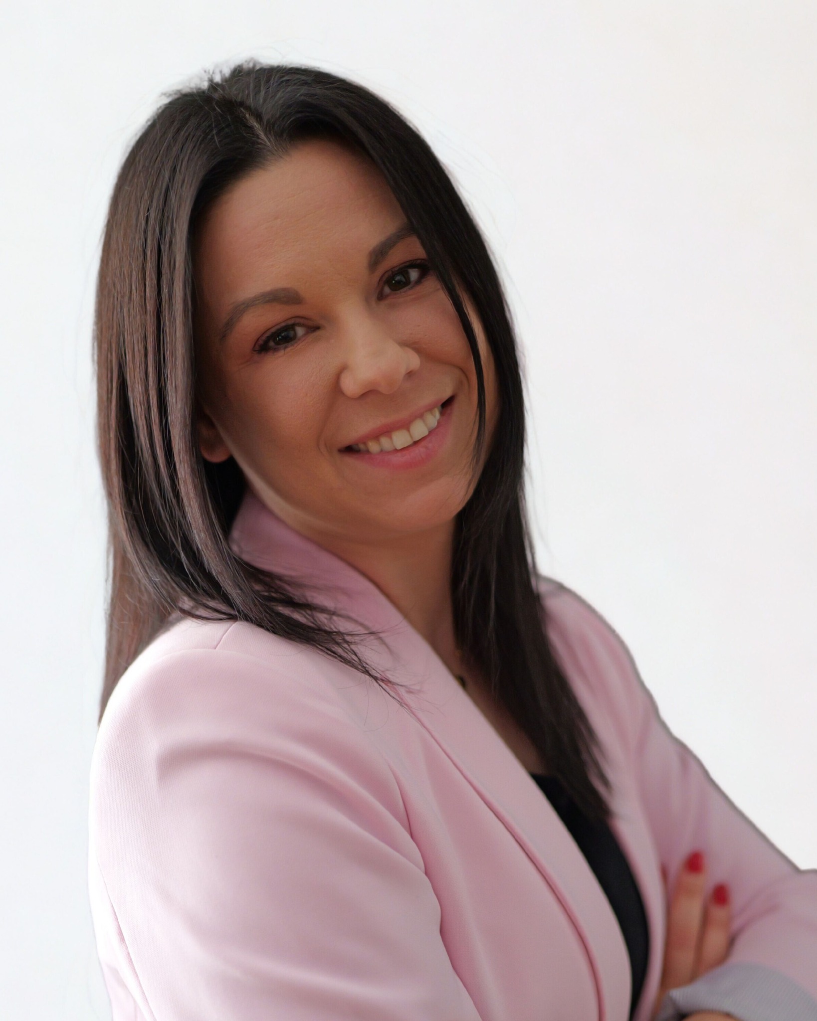 Uśmiechnięta brunetka w różowej marynarce, skrzyżowane ręce, jasne tło. Portret biznesowy, profesjonalny wizerunek doradcy.