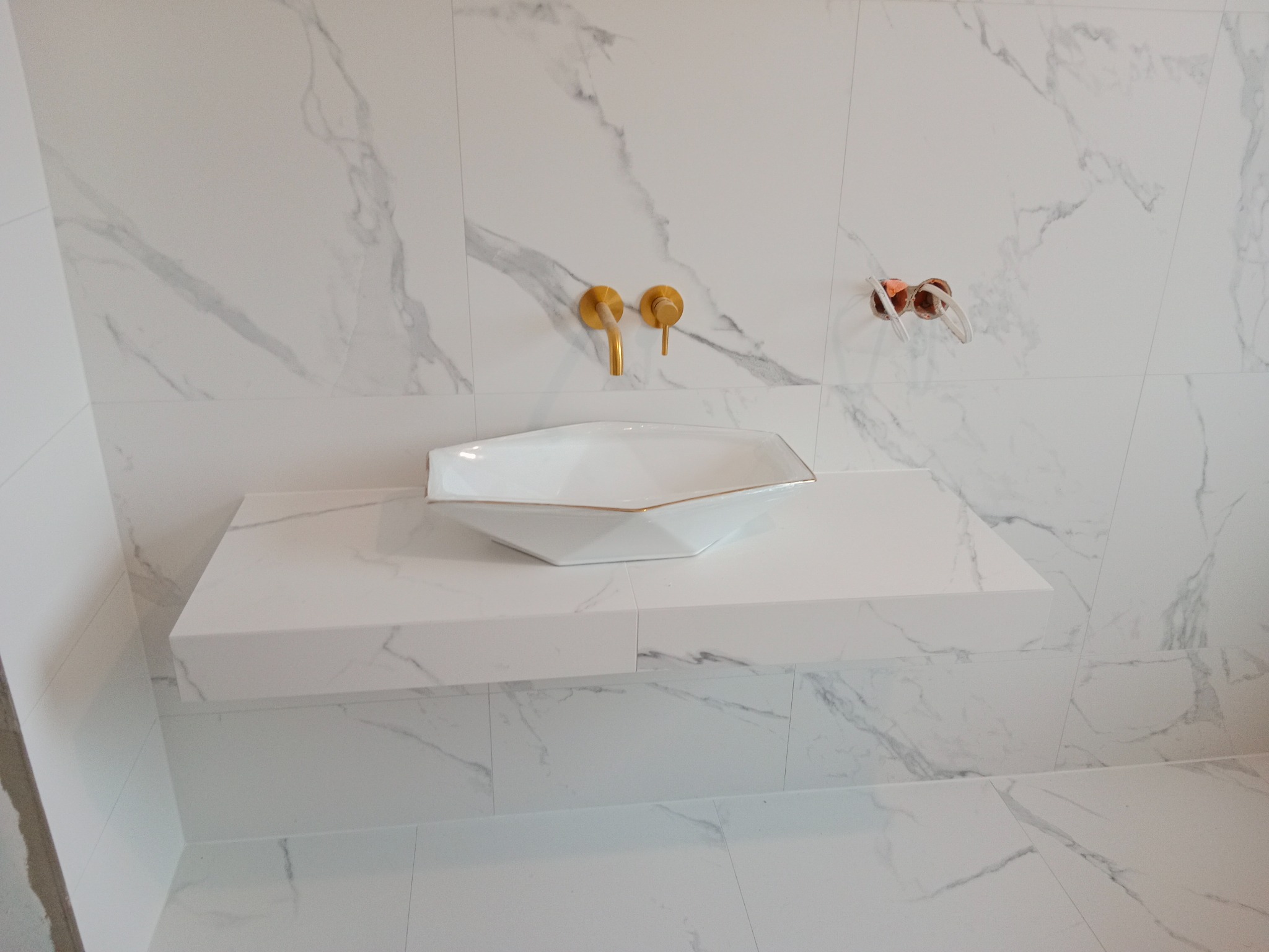 Biała, marmurkowa łazienka z umywalką w kształcie diamentu i złotą armaturą. Widoczne przyłącza instalacji wodnej. Minimalistyczny design, nowoczesne wykończenie.
