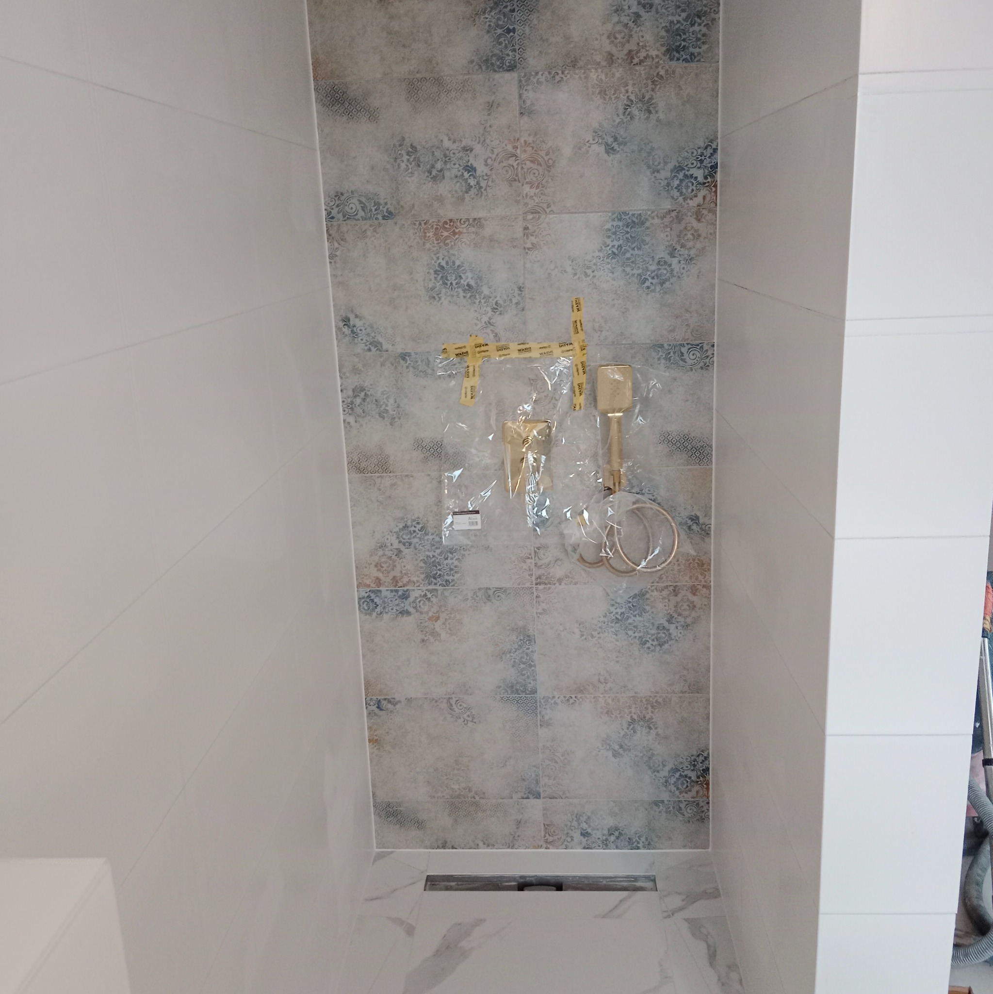 Wykończona ściana w łazience z dekoracyjnymi płytkami i zamontowaną, zabezpieczoną folią, złotą armaturą. Widoczny odpływ liniowy w podłodze.