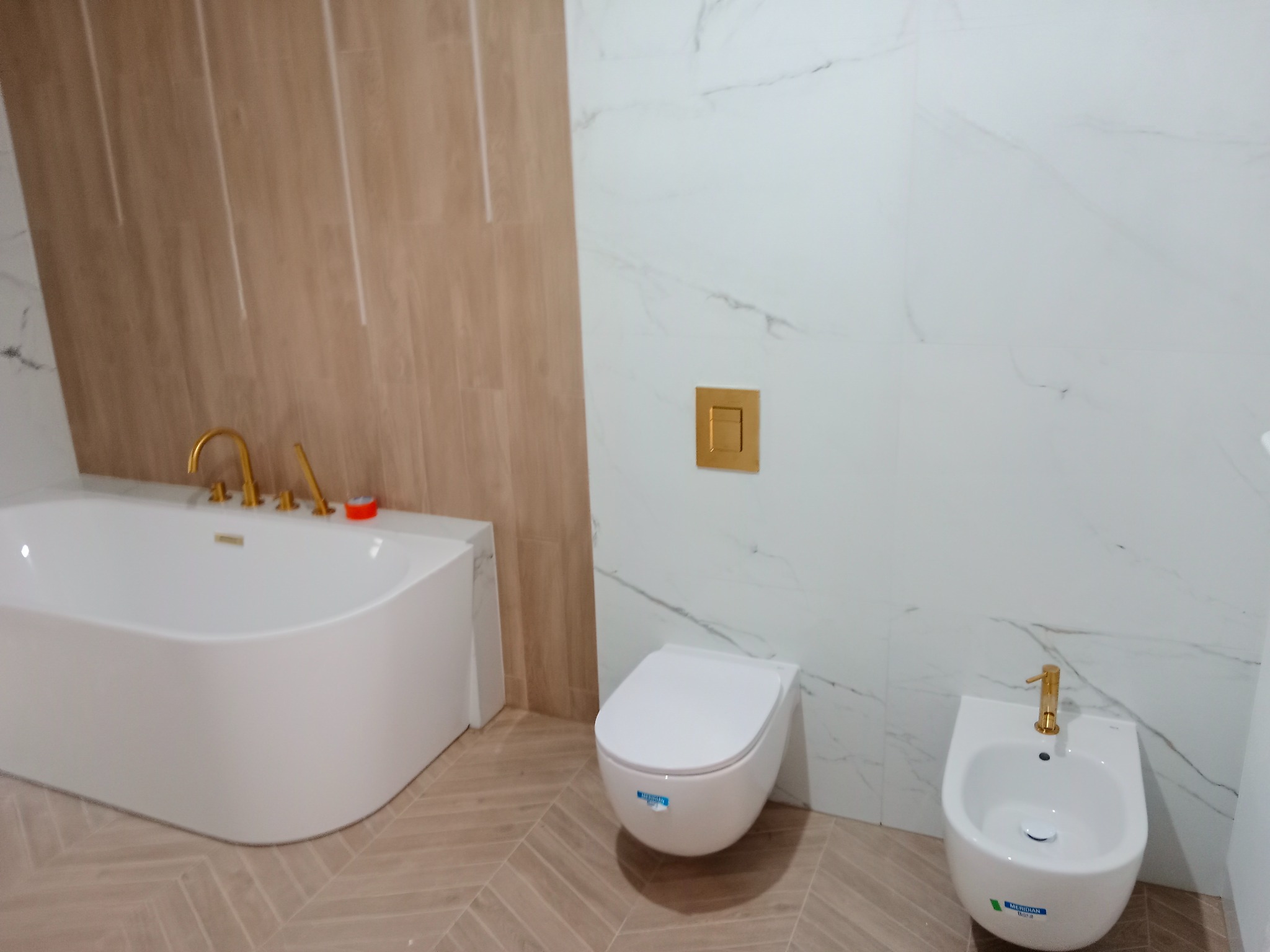 Nowoczesna łazienka z białą wanną, toaletą i bidetem. Złote baterie i armatura. Płytki imitujące drewno na ścianie i podłodze. Marmurowe płytki na ścianie.