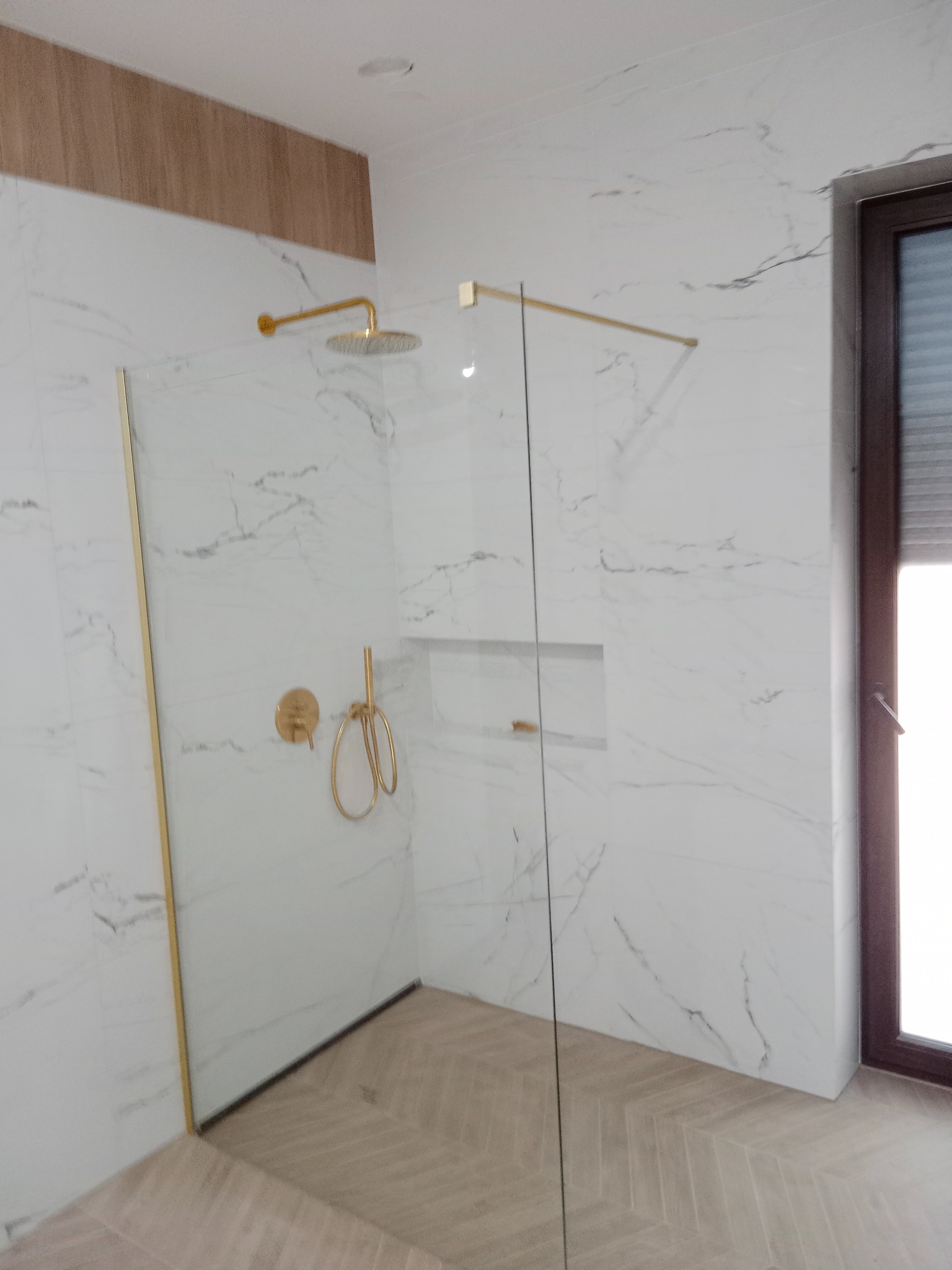 Nowoczesna kabina prysznicowa walk-in z armaturą w kolorze złota, wykończona płytkami imitującymi marmur i podłogą w jodełkę, widok na okno z żaluzjami.