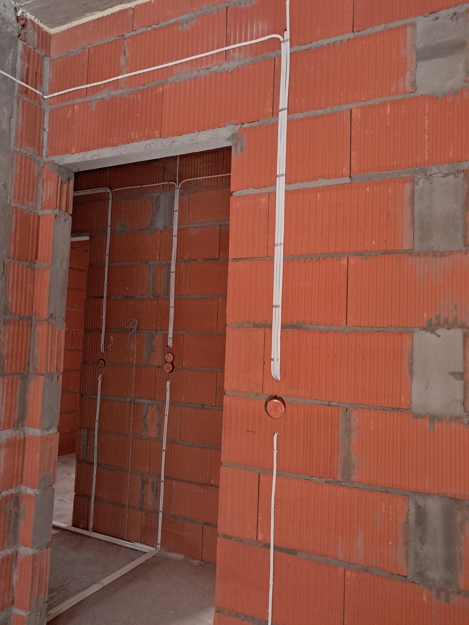 Wewnętrzna instalacja elektryczna w budynku w trakcie budowy. Widoczne kable i puszki w ścianach z czerwonej cegły. Kable prowadzone wzdłuż ścian i nad otworem drzwiowym.