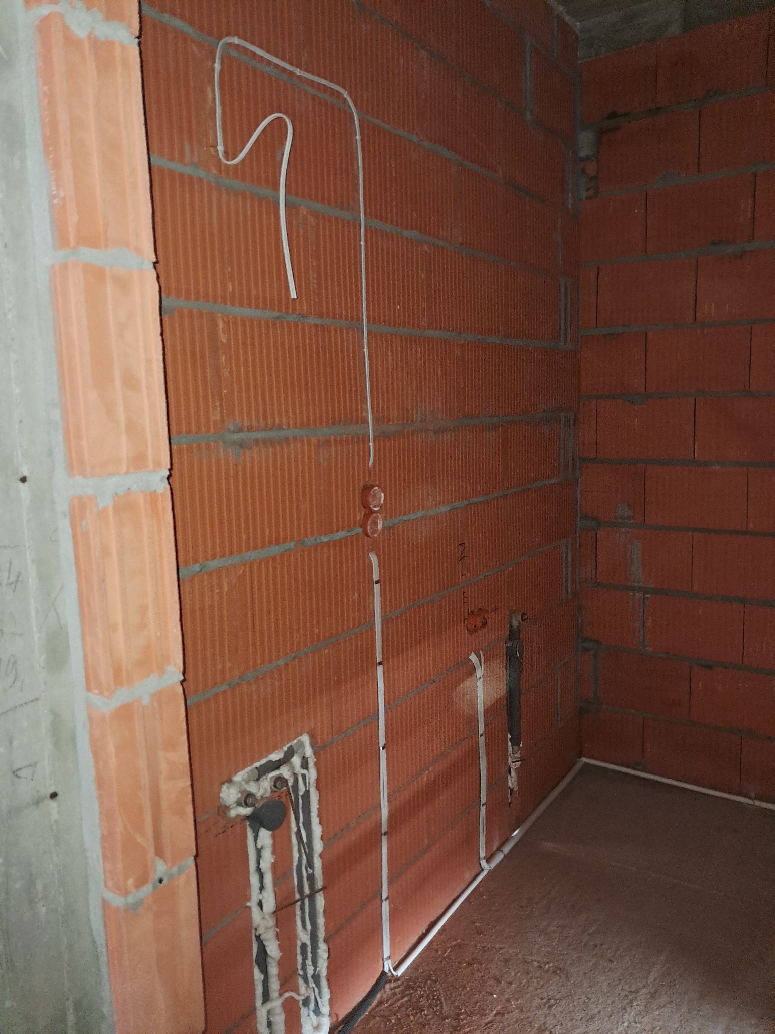 Wewnętrzne ściany z cegły z poprowadzonymi przewodami elektrycznymi w białych rurkach. Widoczne puszki elektryczne i zabezpieczone kable w ścianie. Surowy stan wykończenia.