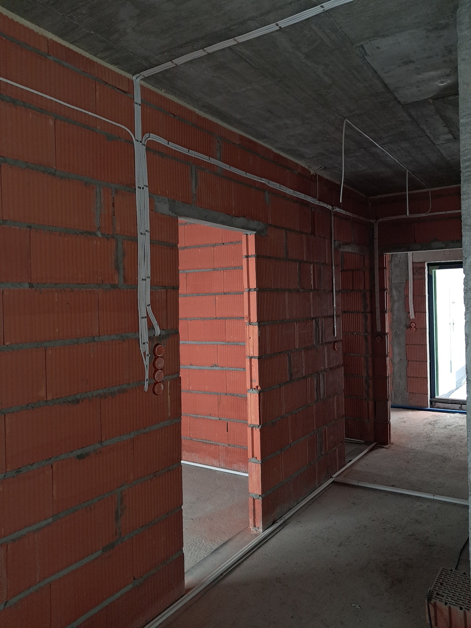 Surowy stan: kable elektryczne prowadzone po ścianach z czerwonej cegły w nowym budynku. Widoczne puszki instalacyjne i peszle. Szare, betonowe sufity.