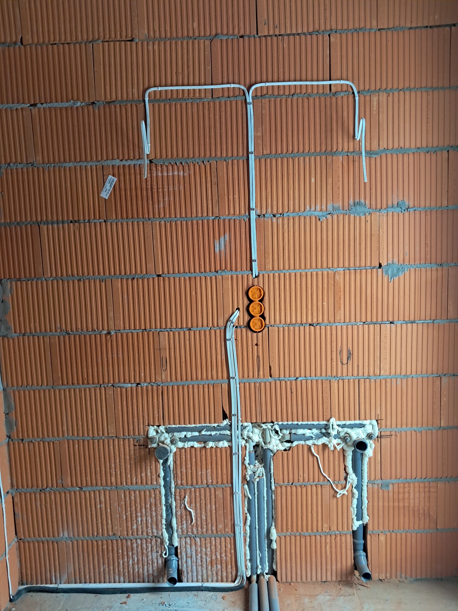 Instalacja elektryczna i hydrauliczna w ścianie z cegły dziurawki. Widoczne kable w peszlach, puszki elektryczne i rury kanalizacyjne zabezpieczone pianką montażową.