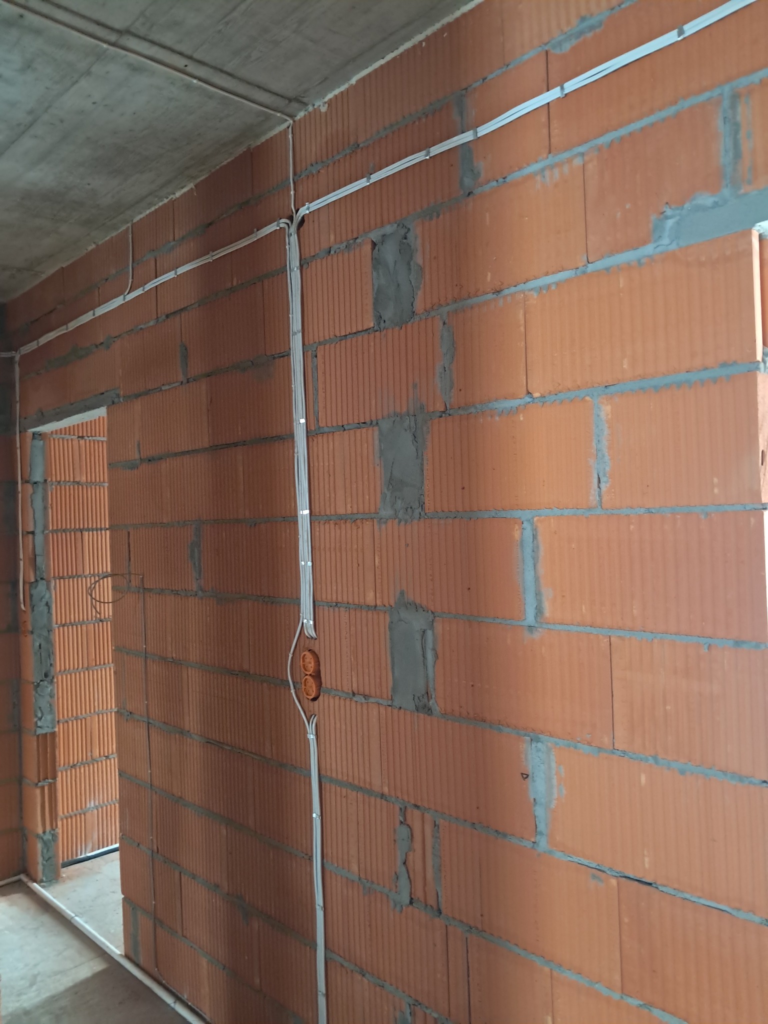 Surowa ściana z cegły z widocznymi przewodami elektrycznymi w białych listwach i pomarańczową puszką. W tle widoczne przejście do innego pomieszczenia.