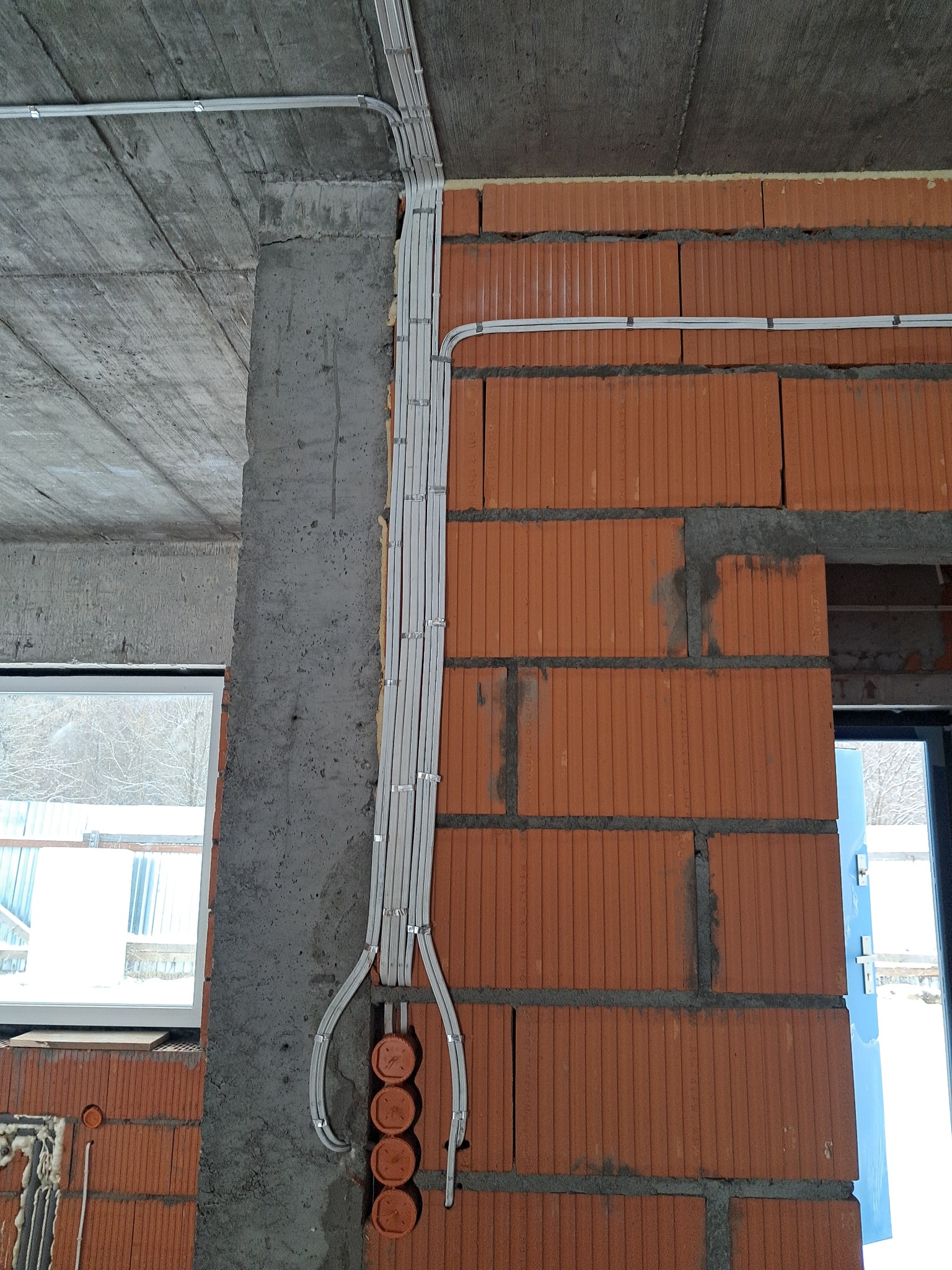 Kable w listwach prowadzących, zamocowane na ścianie z czerwonej cegły i betonowym słupie. Widoczne puszki instalacyjne. Surowy stan wykończenia wnętrza.