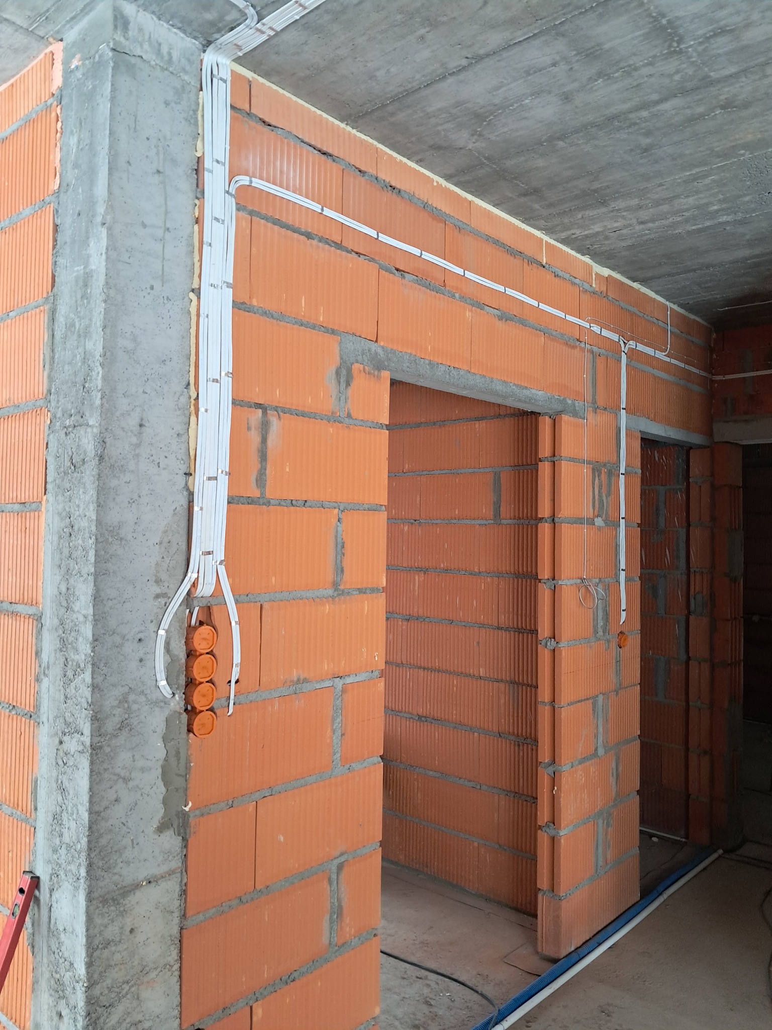 Wewnętrzna instalacja elektryczna w budynku w trakcie budowy. Widoczne kable prowadzone po ścianach z cegły, puszki elektryczne i surowy betonowy strop.