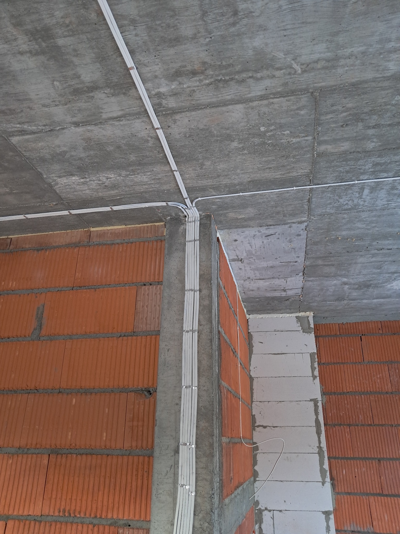 Białe przewody elektryczne ułożone na suficie i ścianie z cegieł, zabezpieczone klamrami. Widok z dołu, surowy betonowy strop i fragment ściany z pustaków.