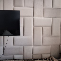Bartosz Jedr - Ściana z beżowych paneli tapicerowanych w geometryczny wzór, z wiszącym telewizorem i fragmentem łóżka. Nowoczesny design wnętrza sypialni.