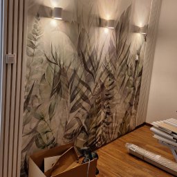 Bartosz Jedr - Ściana z fototapetą w motywie liści, oświetlona trzema kinkietami. Widoczne panele ścienne i podłogowe, karton i narzędzia, sugerujące prace wykończeniowe.