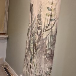 Bartosz Jedr - Fragment ściany z tapetą w motywy roślinne, widoczne listwy przypodłogowe i zabezpieczenia podłogi folią malarską. Tapeta w odcieniach szarości i zieleni na jasnym tle.