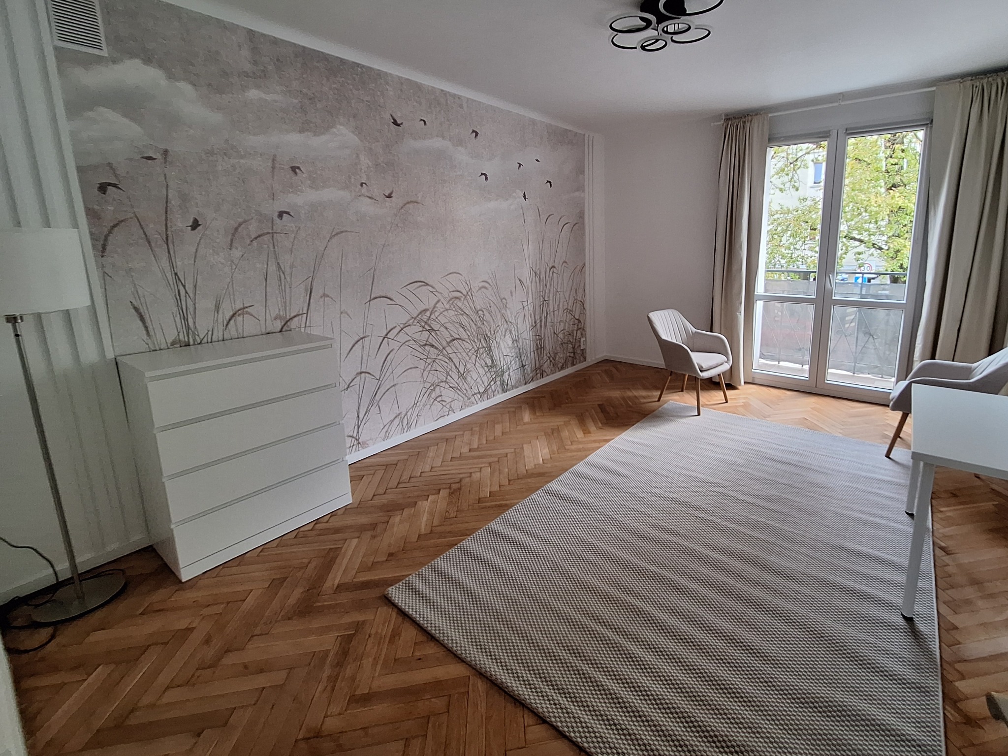 Jasne wnętrze po remoncie: ściana z tapetą w motywy roślinne i ptaki, parkiet w jodełkę, dywan w geometryczny wzór, biała komoda i nowoczesne meble.