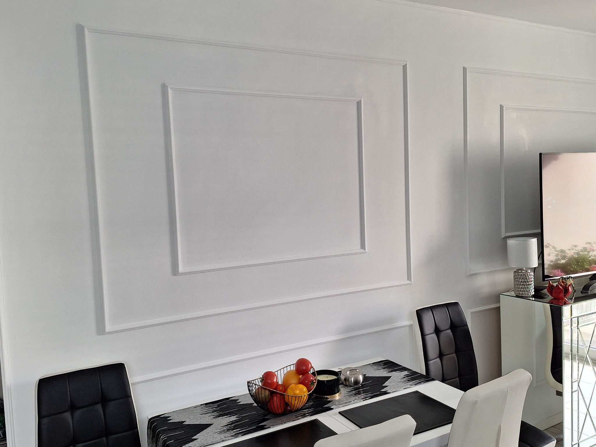 Biała ściana z dekoracyjnymi listwami w kształcie prostokątów, stół z owocami i krzesłami. Minimalistyczne wykończenie wnętrza jadalni w nowoczesnym stylu.
