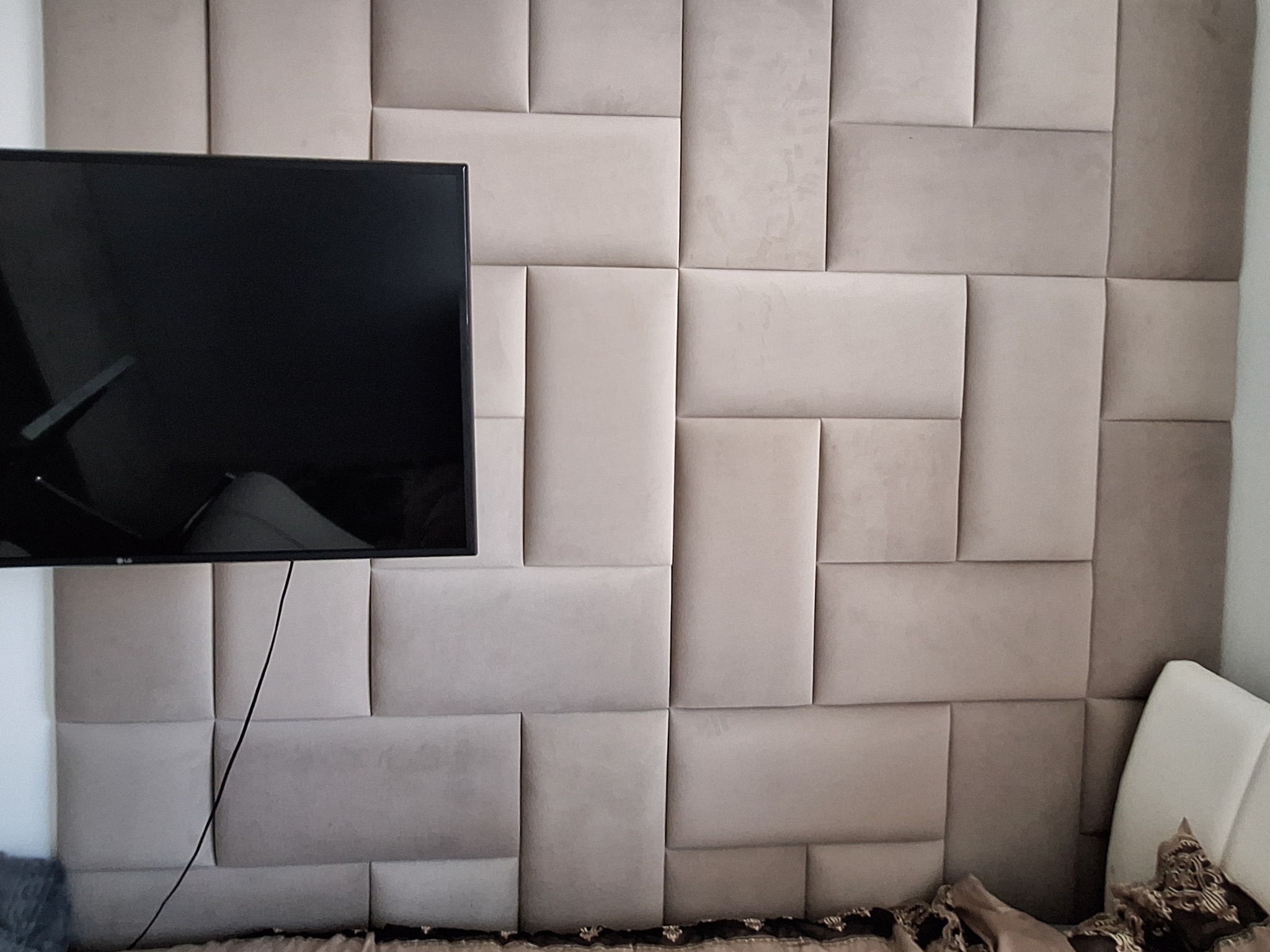 Ściana z beżowych paneli tapicerowanych w geometryczny wzór, z wiszącym telewizorem i fragmentem łóżka. Nowoczesny design wnętrza sypialni.