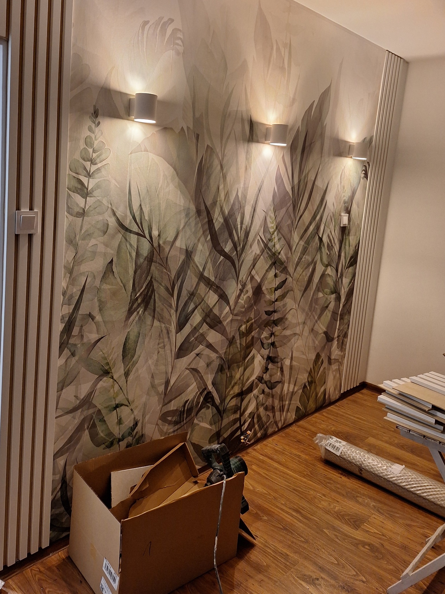 Ściana z fototapetą w motywie liści, oświetlona trzema kinkietami. Widoczne panele ścienne i podłogowe, karton i narzędzia, sugerujące prace wykończeniowe.
