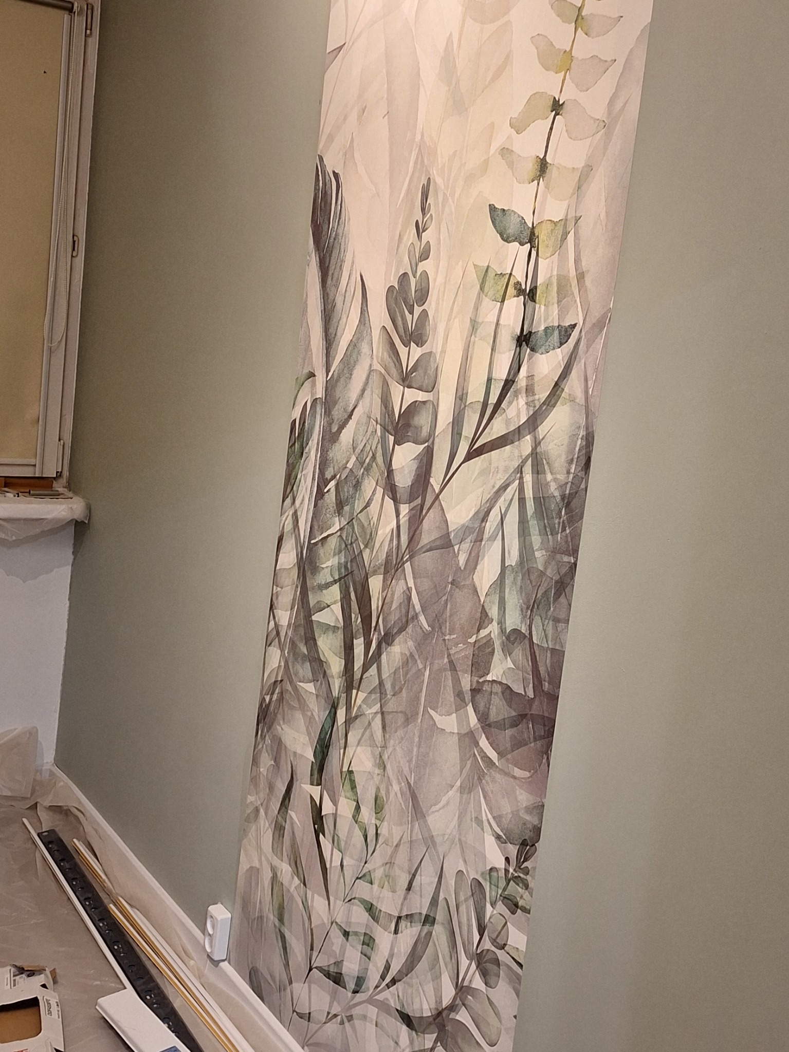 Fragment ściany z tapetą w motywy roślinne, widoczne listwy przypodłogowe i zabezpieczenia podłogi folią malarską. Tapeta w odcieniach szarości i zieleni na jasnym tle.