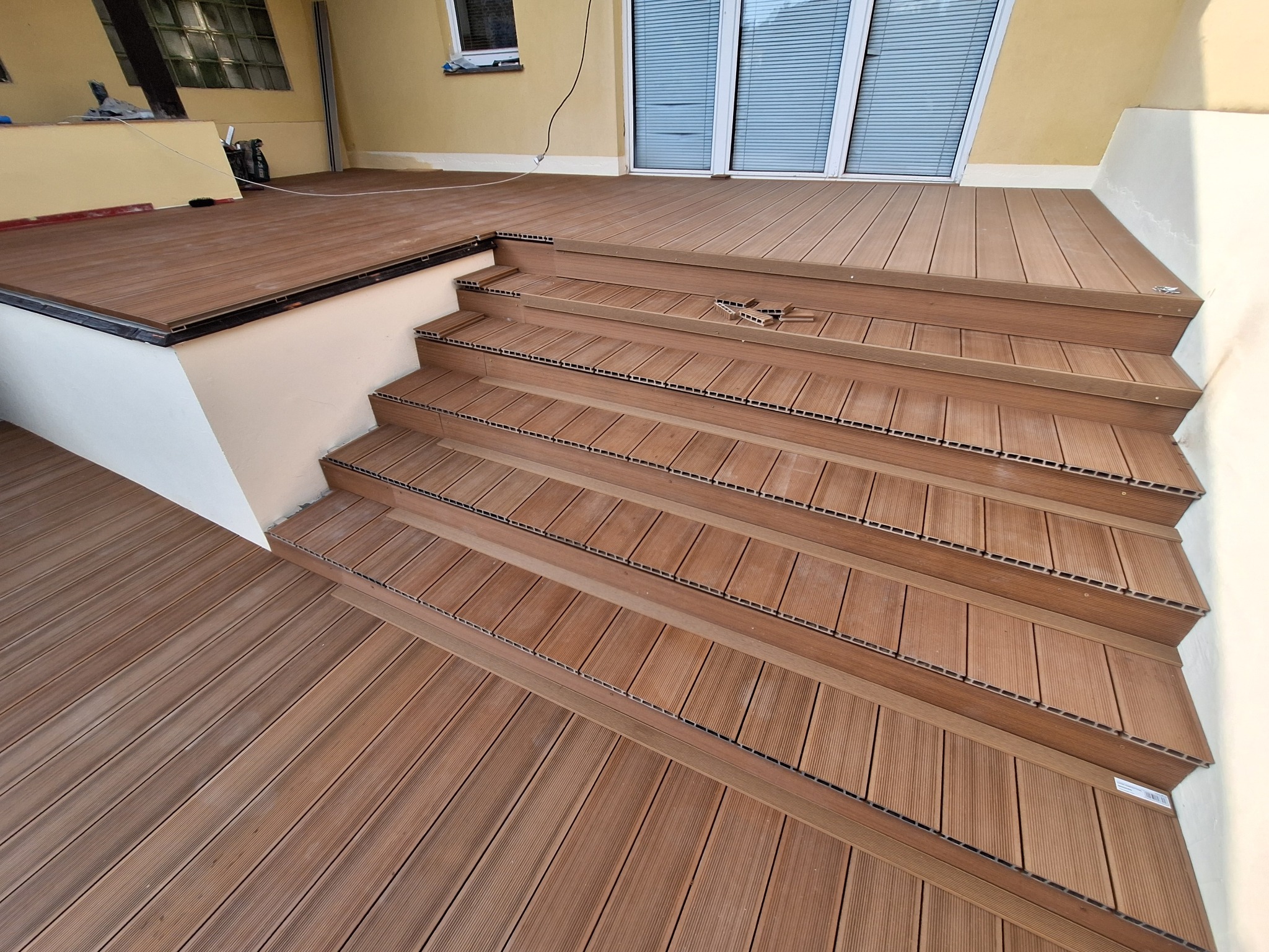 Wykończony taras z kompozytowych desek w odcieniach brązu, prowadzący schodami do drzwi balkonowych. Widoczne detale montażu i struktura desek.