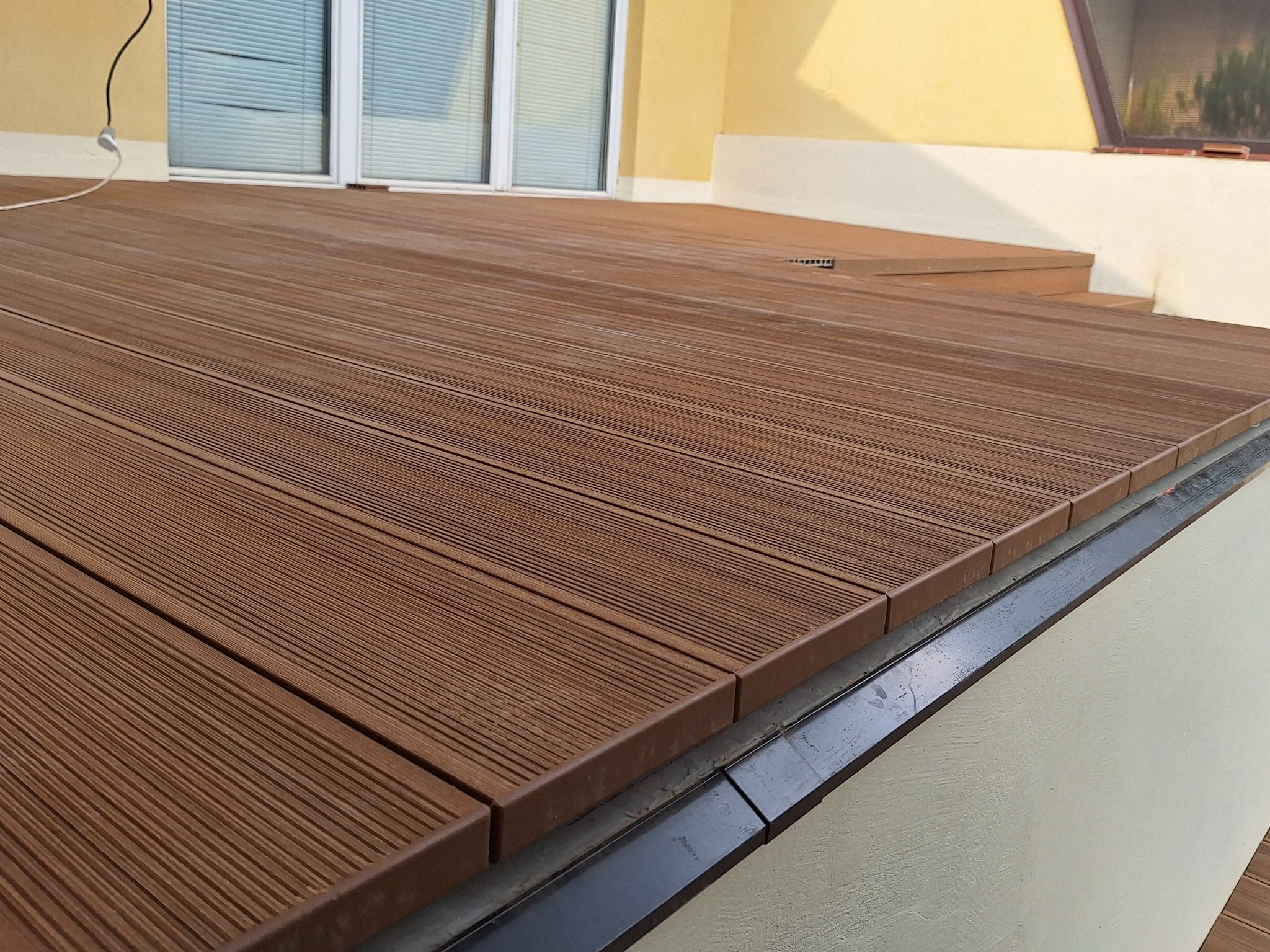 Nowoczesny taras z desek kompozytowych w odcieniach brązu, z widocznym odpływem liniowym i fragmentem elewacji w ciepłych barwach. Estetyczne wykończenie.