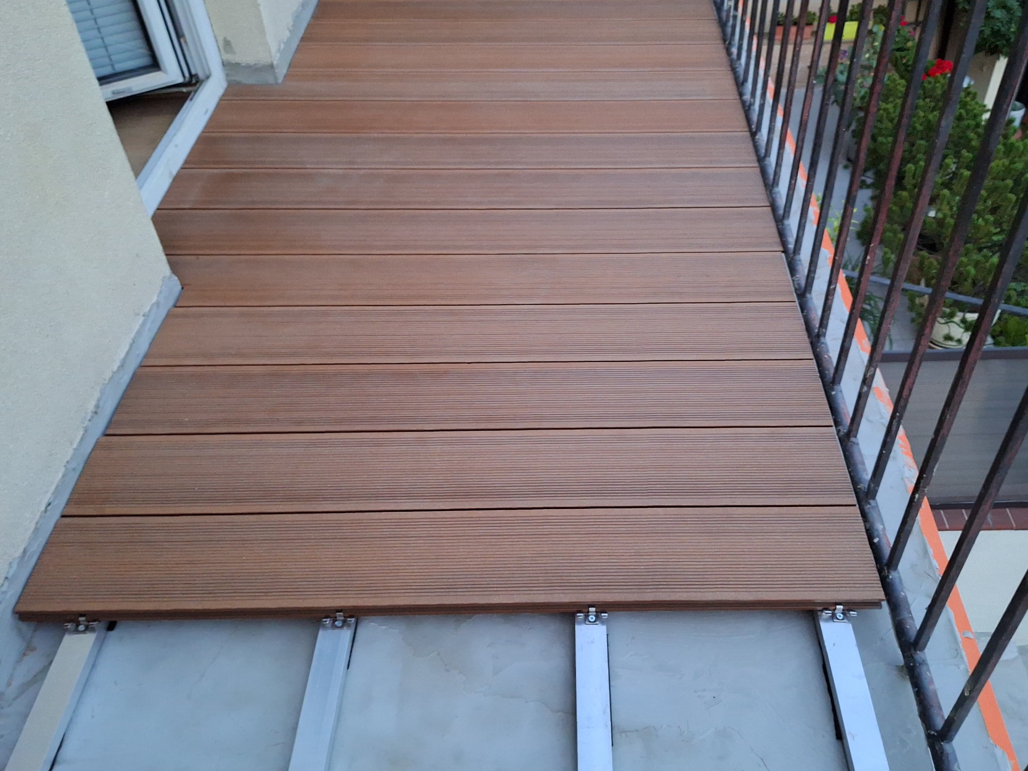 Balkon z brązową podłogą z desek kompozytowych, widoczne aluminiowe legary i mocowania. Balustrada z czarnych prętów. W tle rośliny w donicach.