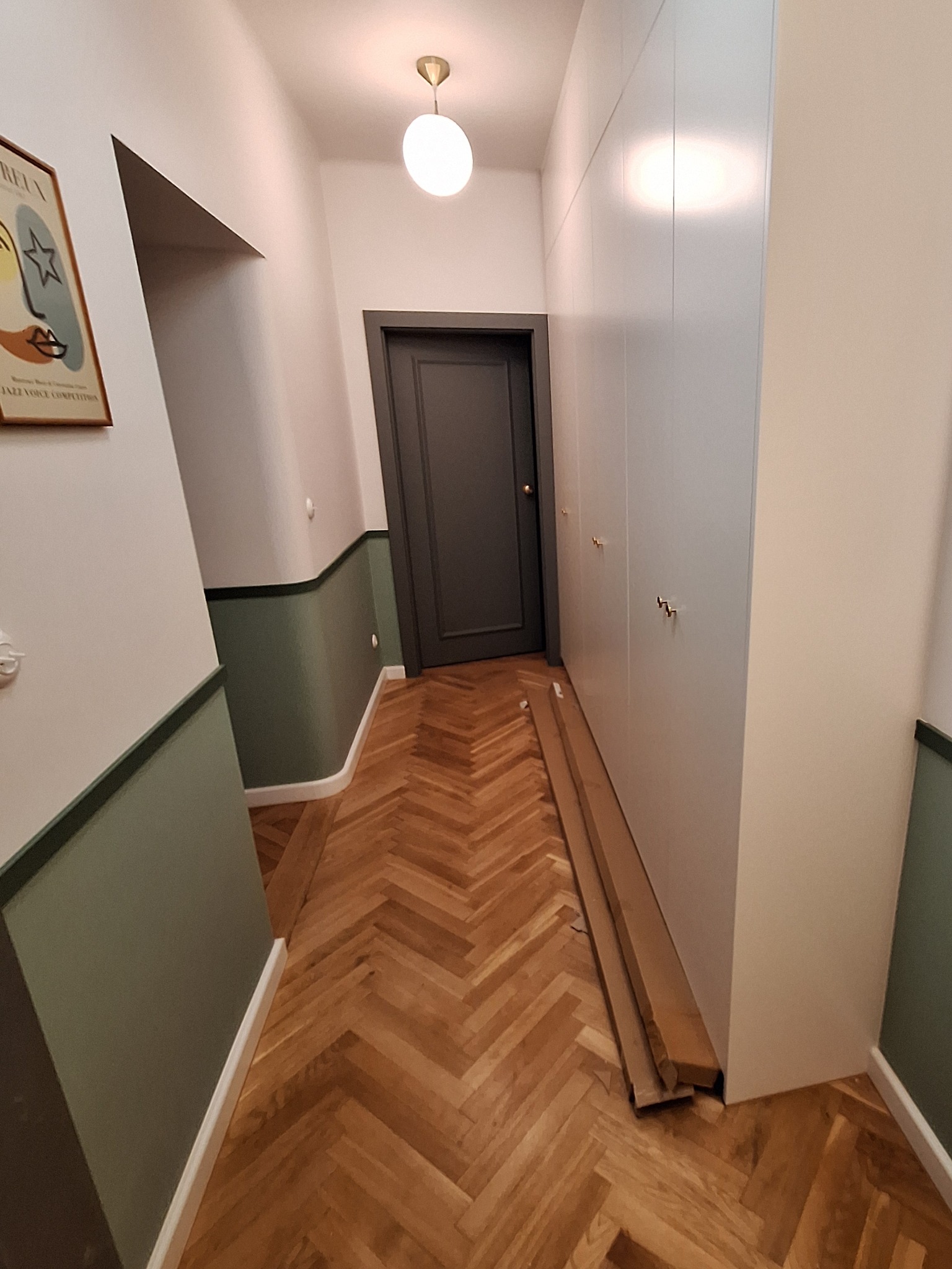Korytarz po remoncie: parkiet w jodełkę, ściany z lamperią w odcieniach zieleni, szara stolarka drzwiowa i biała szafa w zabudowie z delikatnymi, złotymi uchwytami.