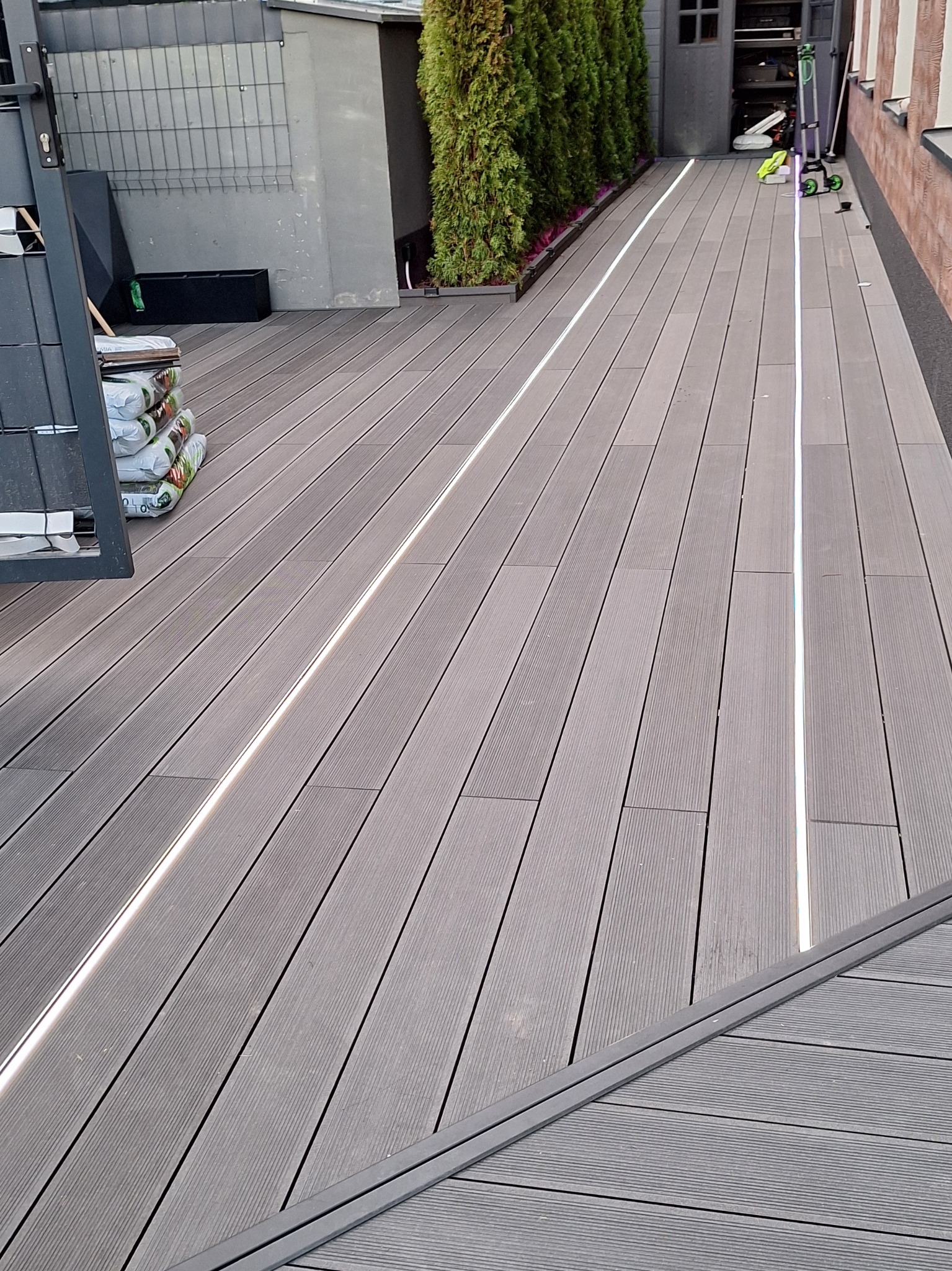 Szara deska kompozytowa z wbudowanym oświetleniem LED na tarasie. W tle roślinność i elementy architektury ogrodowej. Widok z góry, ukośny kadr.