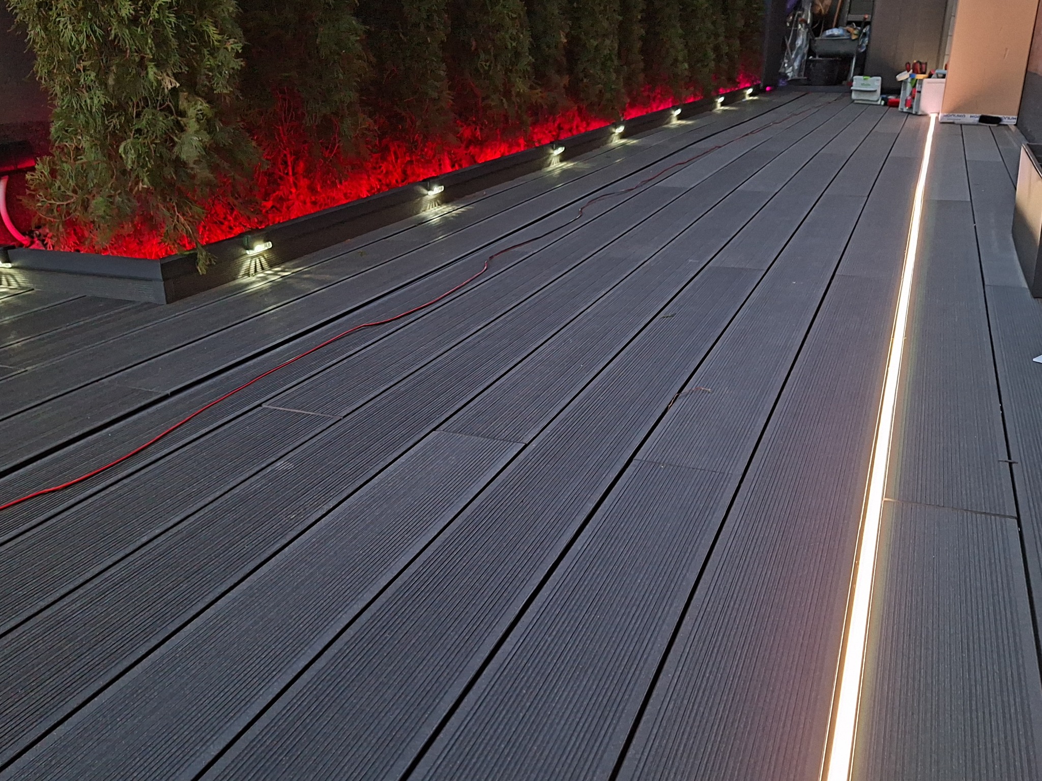Nowoczesny taras z desek kompozytowych oświetlony liniowym światłem LED i czerwonymi lampkami przy roślinach, tworzącymi nastrojowy klimat.