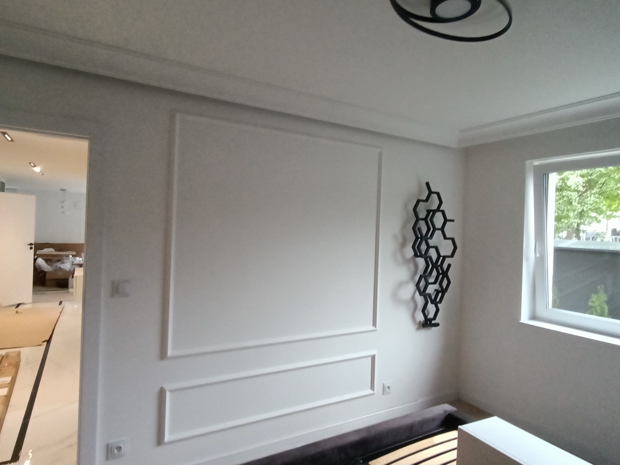 Białe ściany z dekoracyjnymi listwami i czarną ozdobą w kształcie plastra miodu. Widok na okno i fragment wnętrza w tle. Minimalistyczny design.